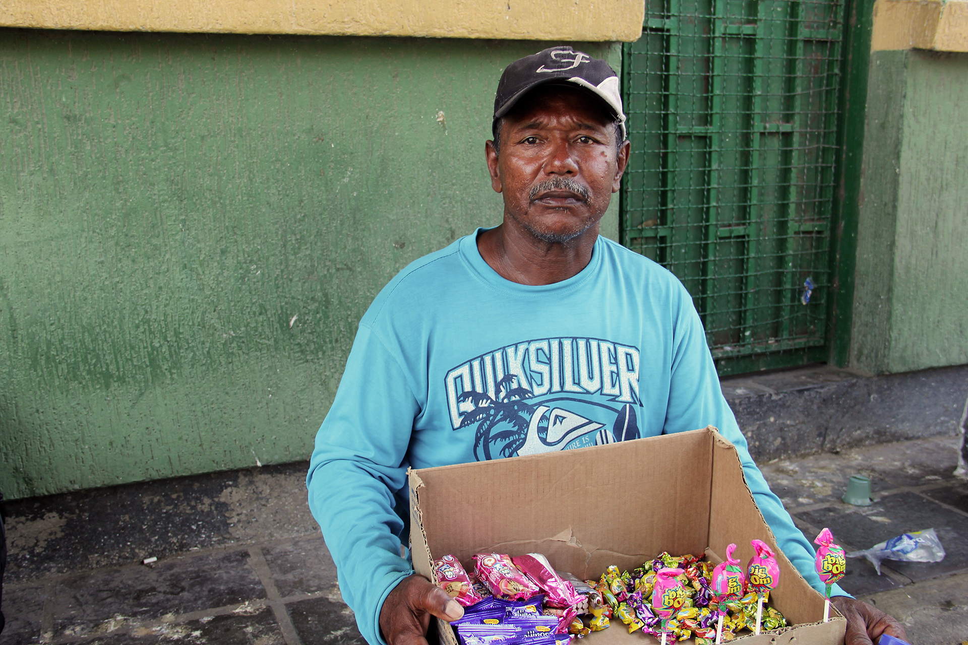 A 48 year old man stands near a colorful wall holding a box of small goods for sale