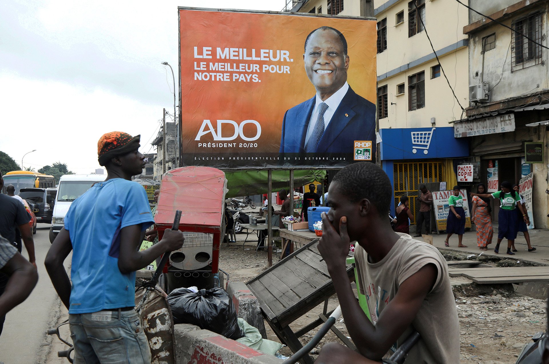 People look on in front of a campaign billboard of presidential candidate Alassane Ouattara of the ruling RHDP coalition in Adjame district, Abidjan, Ivory Coast on 15 October, 2020.