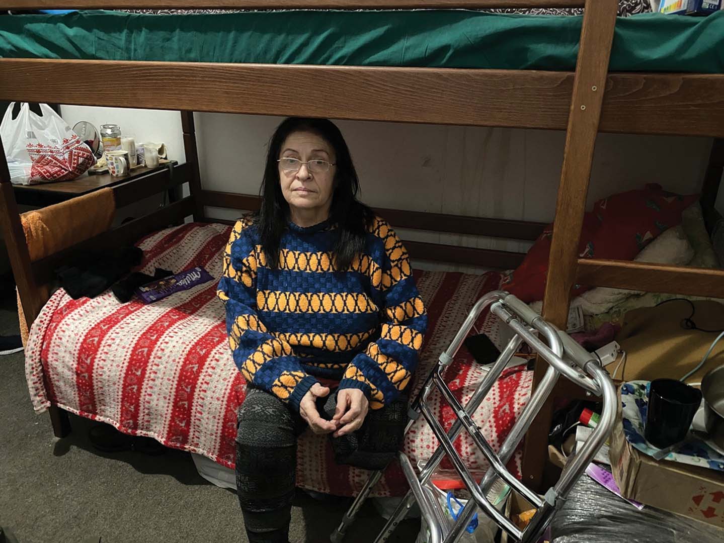 Liudmyla Daniuk, 60, pictured in her room her room in a seminary in Lviv, Ukraine in January 2023, where she is living after returning from Slovakia. She's sitting at the edge of the bed with her hands on her lap. To her left side is a walker.