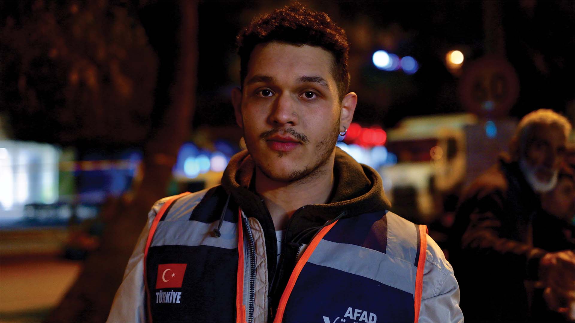 It's nighttime. Volunteer Barış Bilir stands near the site of a collapsed building in Adana. 