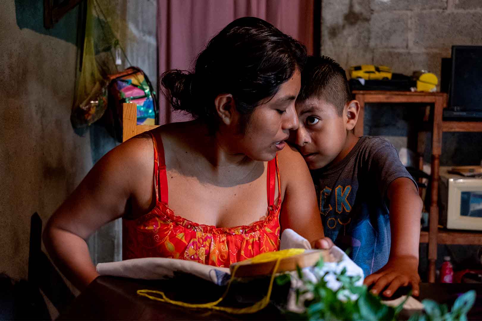 Medium shot of a mother and a son. The mother is holding a book on her lap her face is turned to her left shoulder where her son rests his cheek as he looks up to her. 