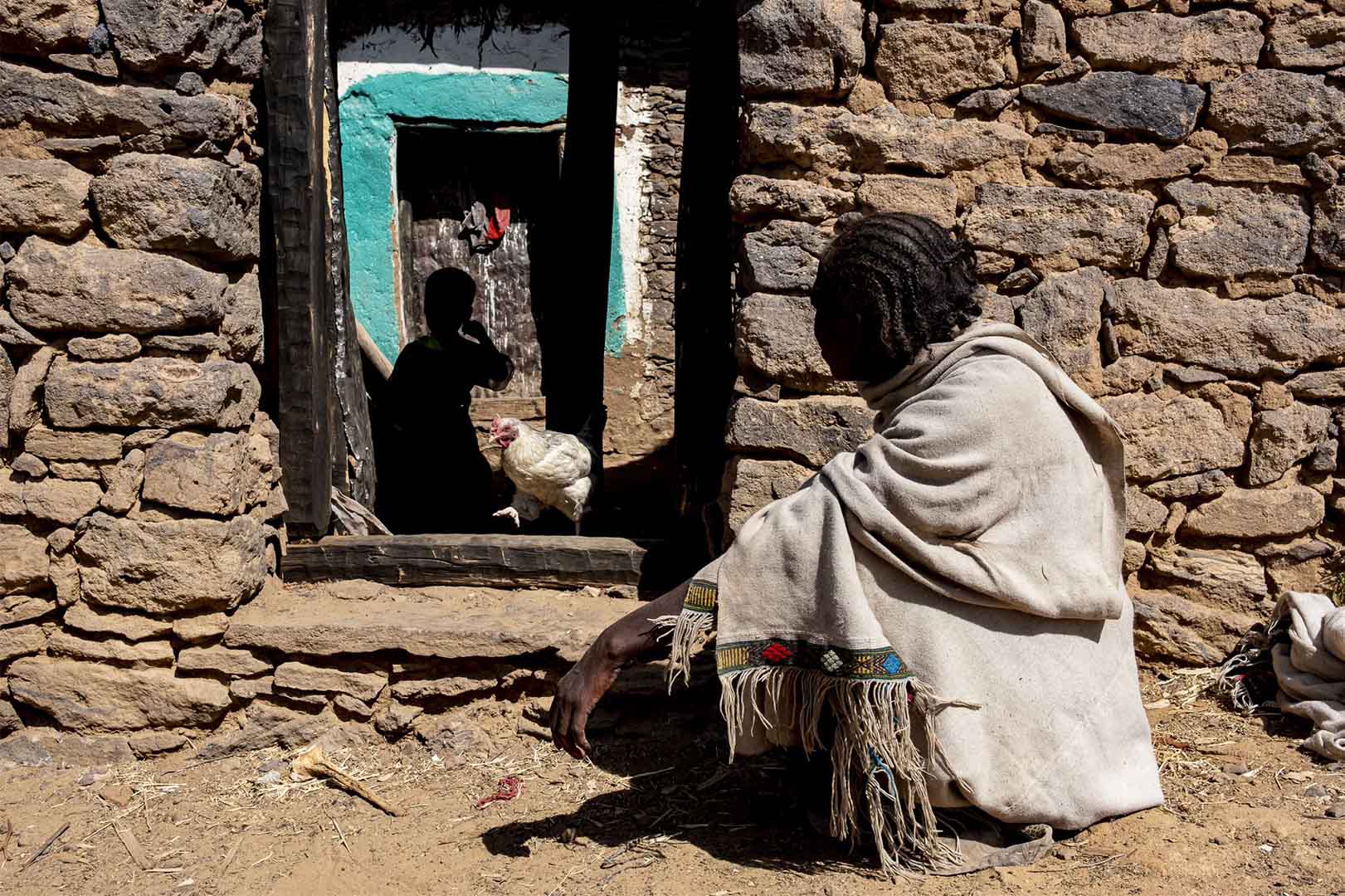 A woman is crouched outside her home by the frame of her door. Inside the frame we see the silhouette of a child.