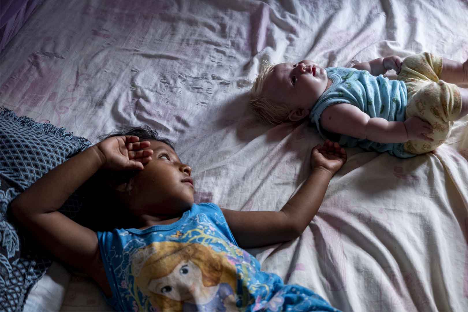 A toddler and a baby lie in a bed. The toddler is reaching out gently to the baby with their left hand.