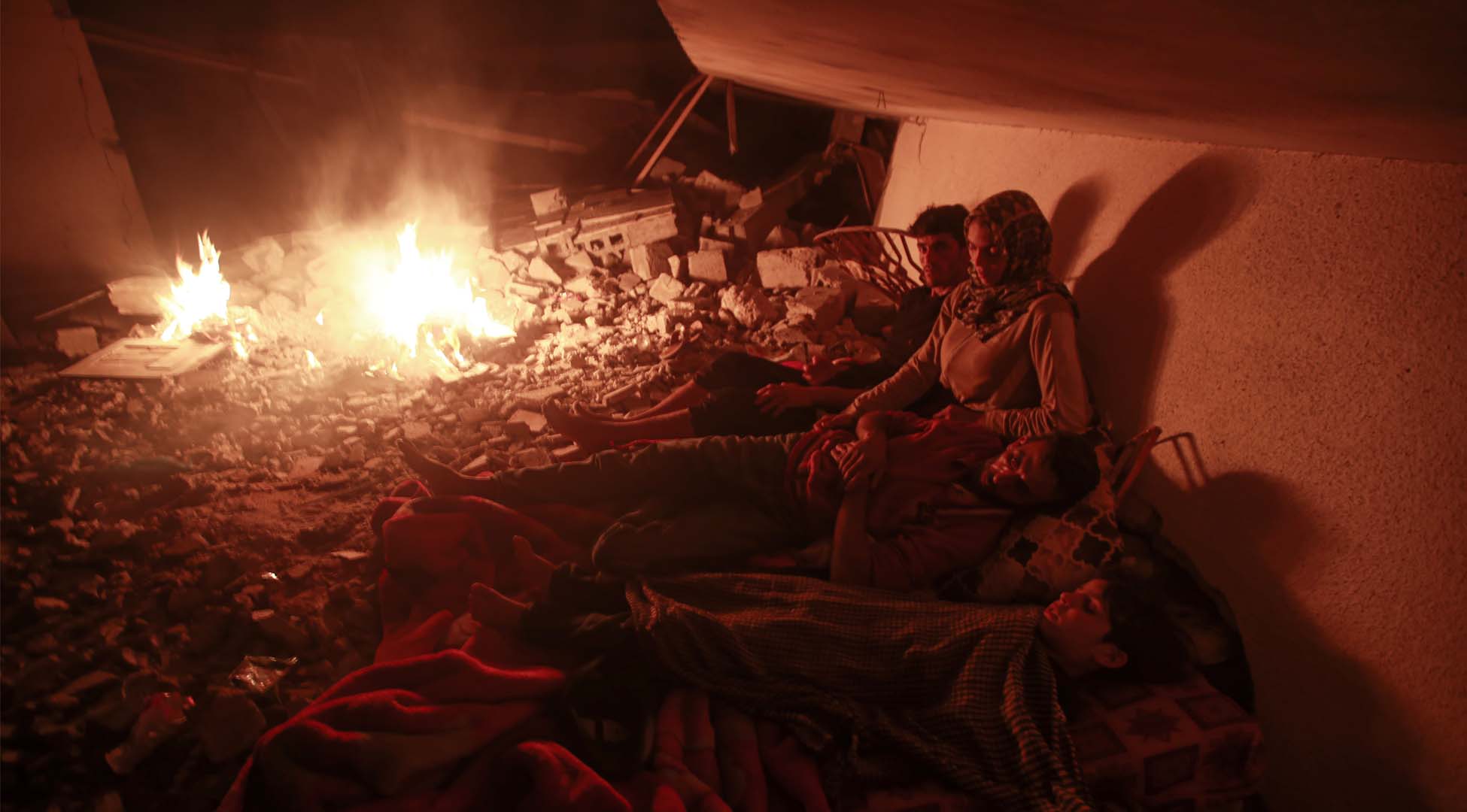 Pictured here is a family that has been left homeless by the bombing, and is taking shelter near the ruins of their destroyed home. There are two men, one woman and one boy. One of the men and the boy are laying on top of what looks to be a mattress which has been placed on top of rubble. The woman and other man are sitting on the rubble. It is night time, and the detroyed home is illuminated by a campfire that is to the right of the family. 