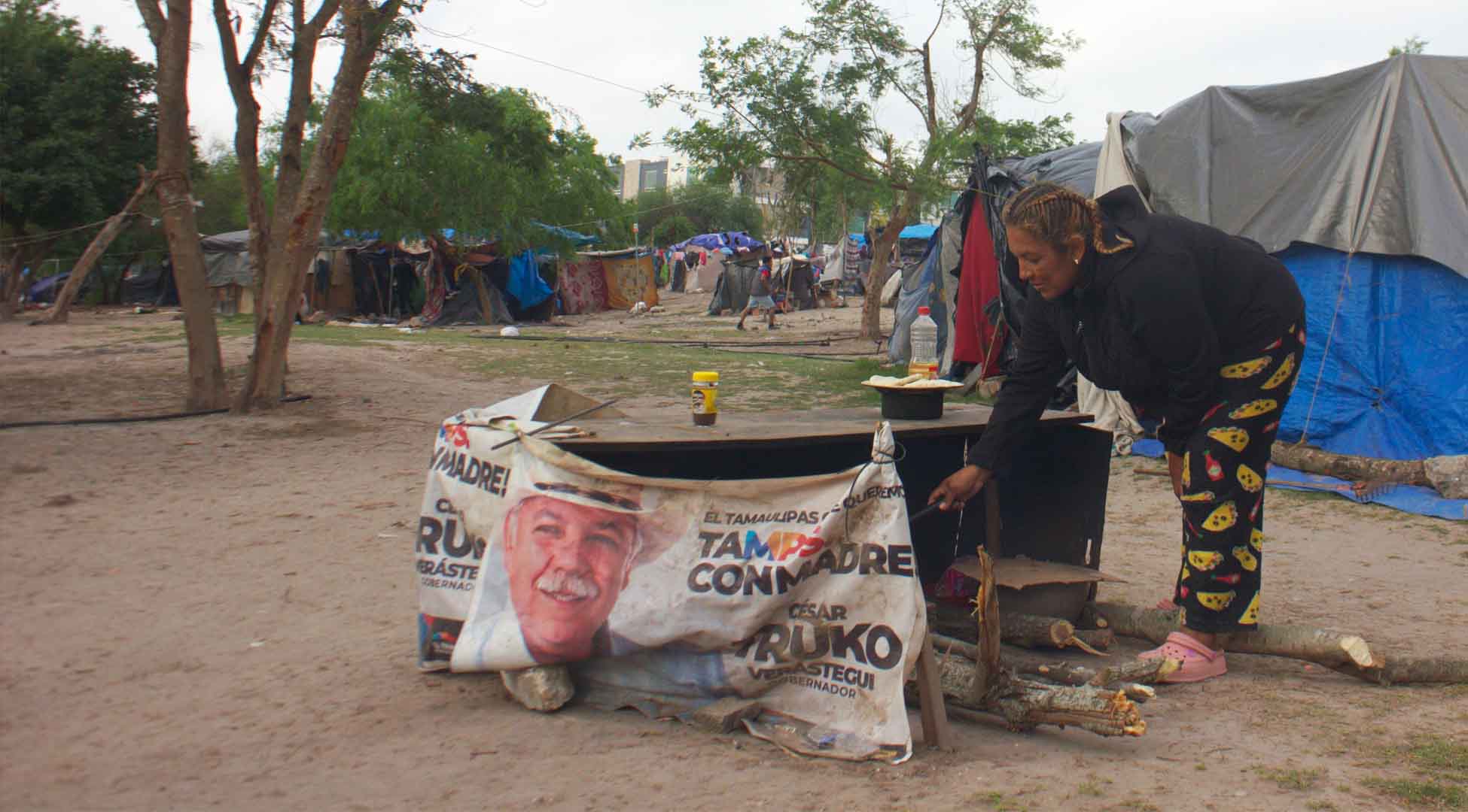 At the center of the image is a woman bending at the waist cooking in a makeshift fire. Behind her are makeshift tents. There are trees in the background. 