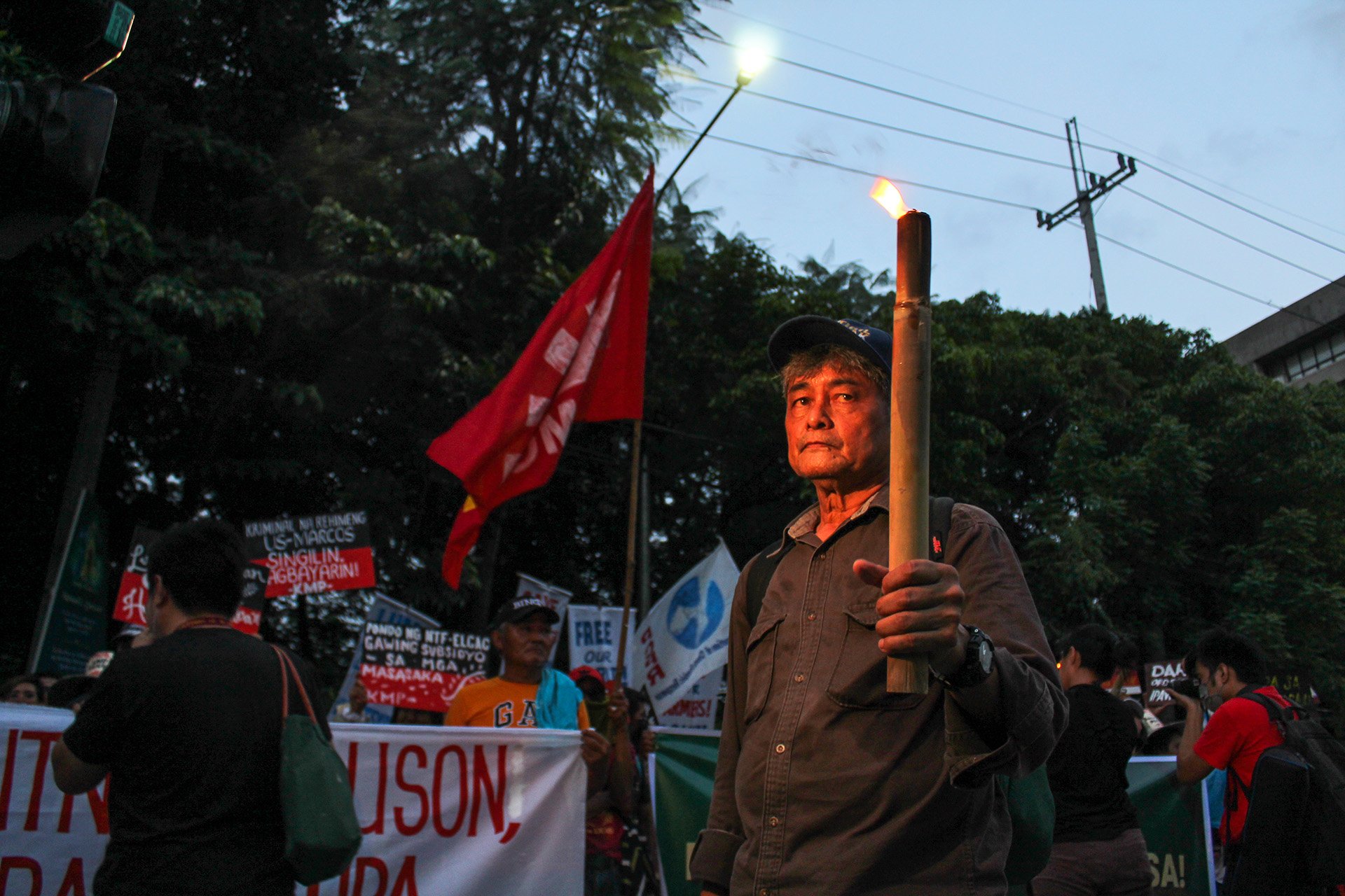 Dusk at a protest, a man holds a lit torch and looks at the camera.