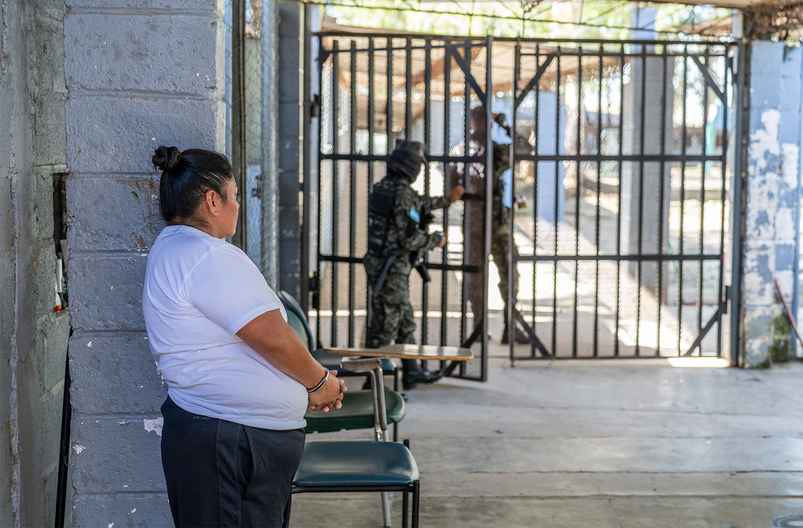 Viena Rosana Sances Martinez, 38, an inmate, stands to the left of the frame leaning against a wall. Her hands are handcuffed. She wears a white shirt and black pants. In the background we see two guards and a gate.