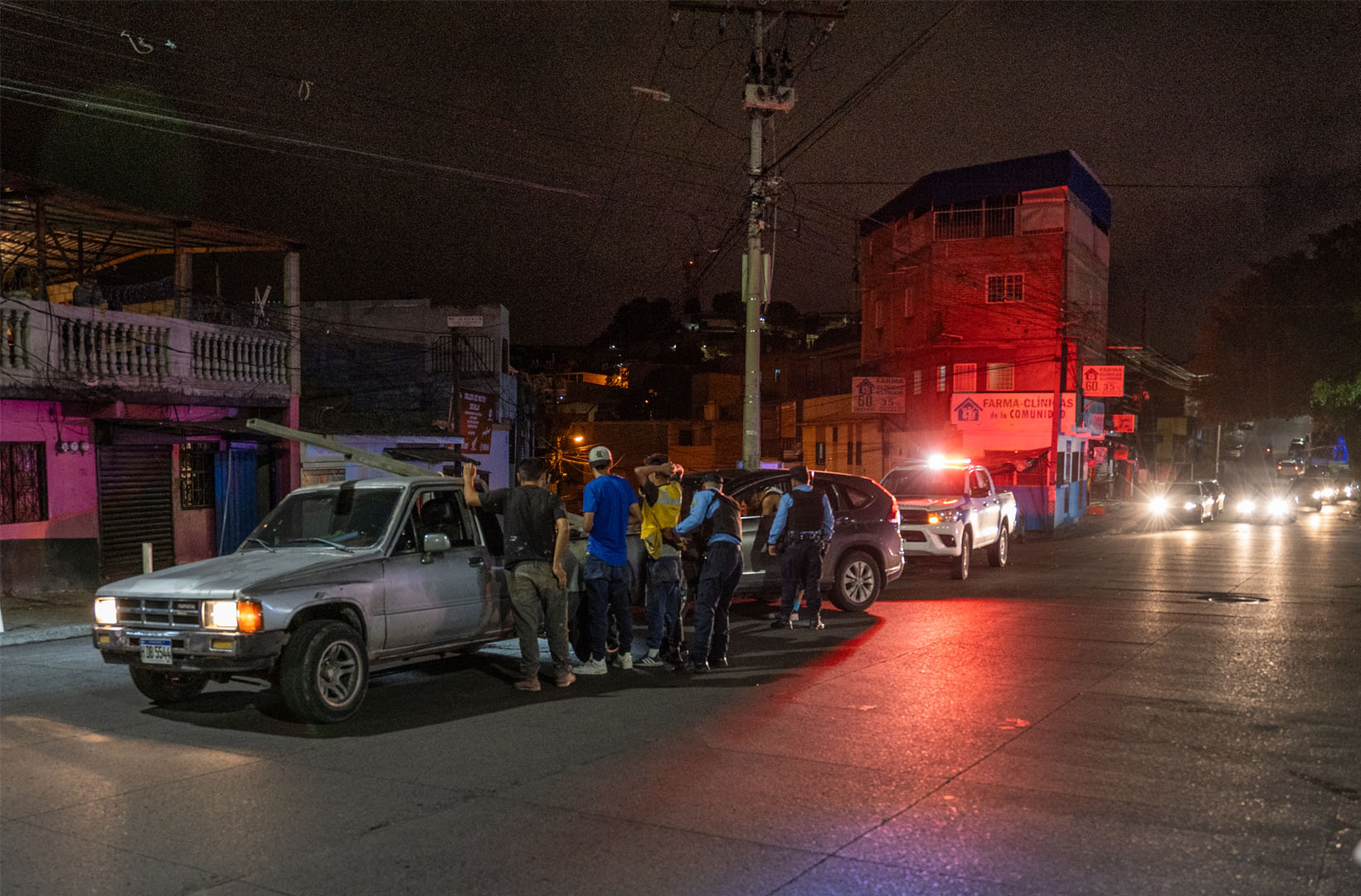 This picture is taken at night. We can see two cars parked on the right side of a road, behind them is a police car. Stood against the first car are three individuals with two police officers searching them.