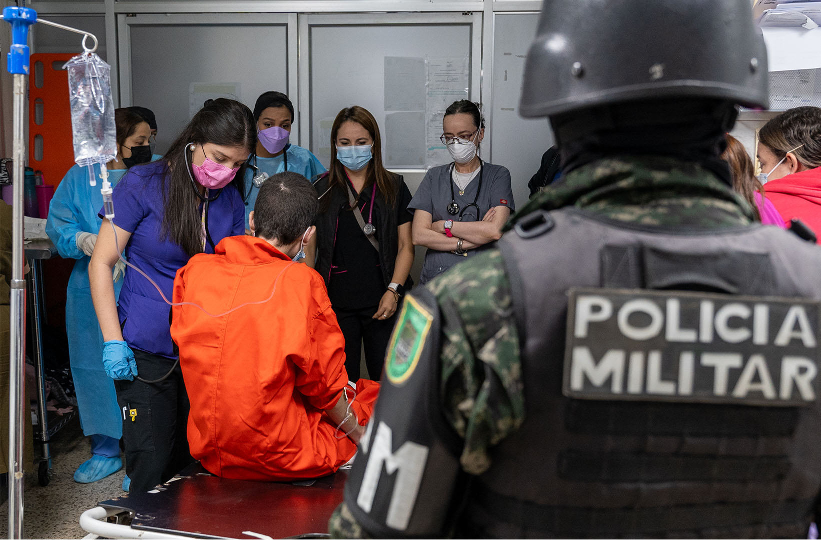 This picture is taken from within a hospital. It's an over the shoulder shot of a military police officer. In front of him is Emerson Roney Diaz (21), a member of the Barrio 18, receiving treatment of a gunshot wound obtained in prison. We see his back as he is treated by a doctor in dark blue scrubs as others look on.