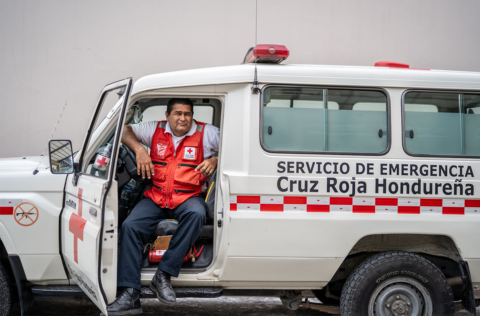 This is a picture of alex Martínez (50), a volunteer paramedic who has worked for 27 years in the Honduran Red Cross, waiting for his colleague to return from turning over a patient as he sits in the front seat of an ambulance. The door is open and he is seated facing the outside of the car.