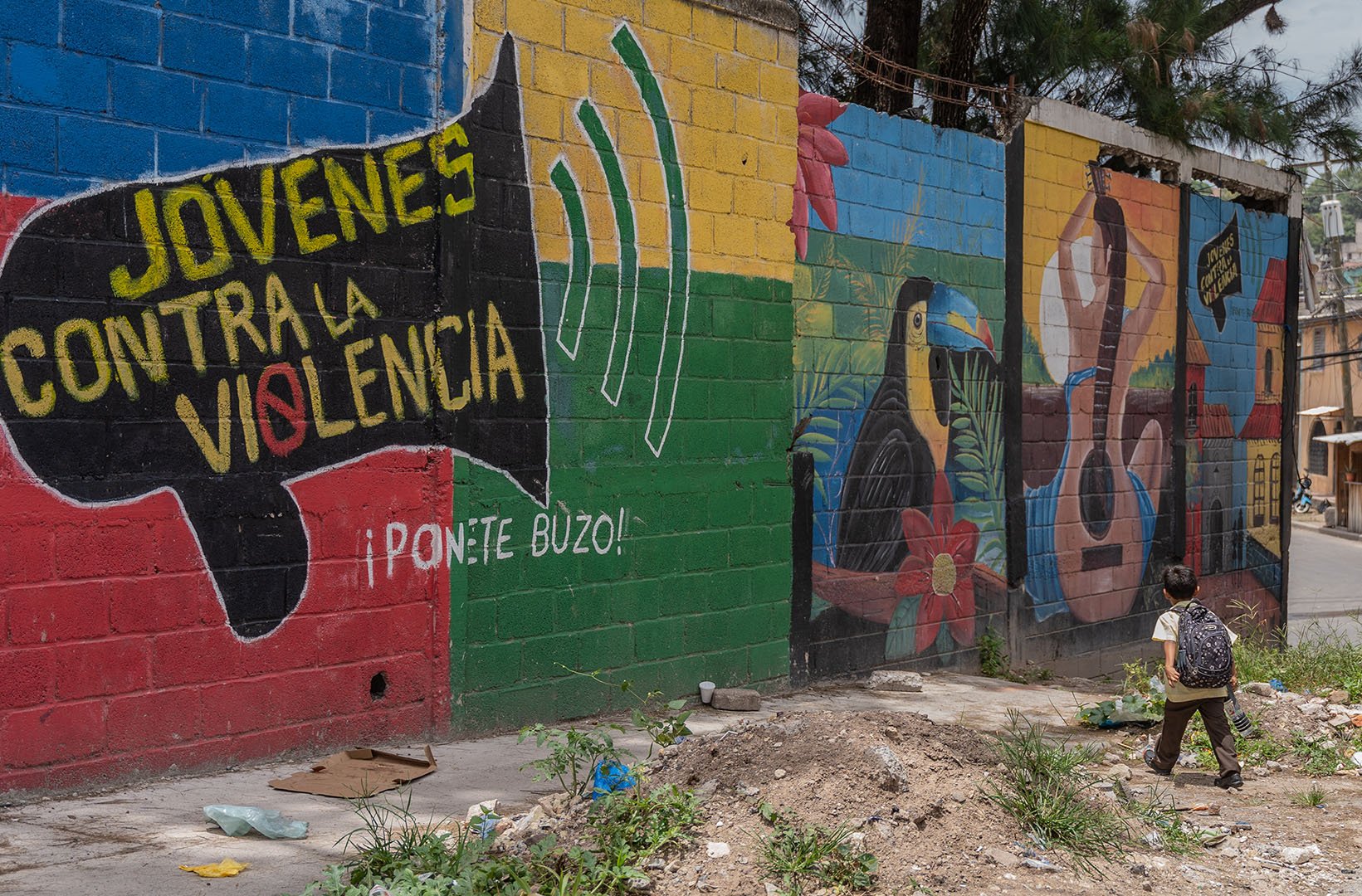 This is a picture of a large mural that says "Jovenes contra la violencia"(Youth against violence). These words are painted into the drawing of a loudspeaker over a backgrouns of blue, yellow, red and green boxes. At the bottom right we see a young child walking away from the camera. This mural is part of the programme ‘Decorando mi barrio’ (Decorating my Neighborhood) which seeks to paint over the gang graffitis.