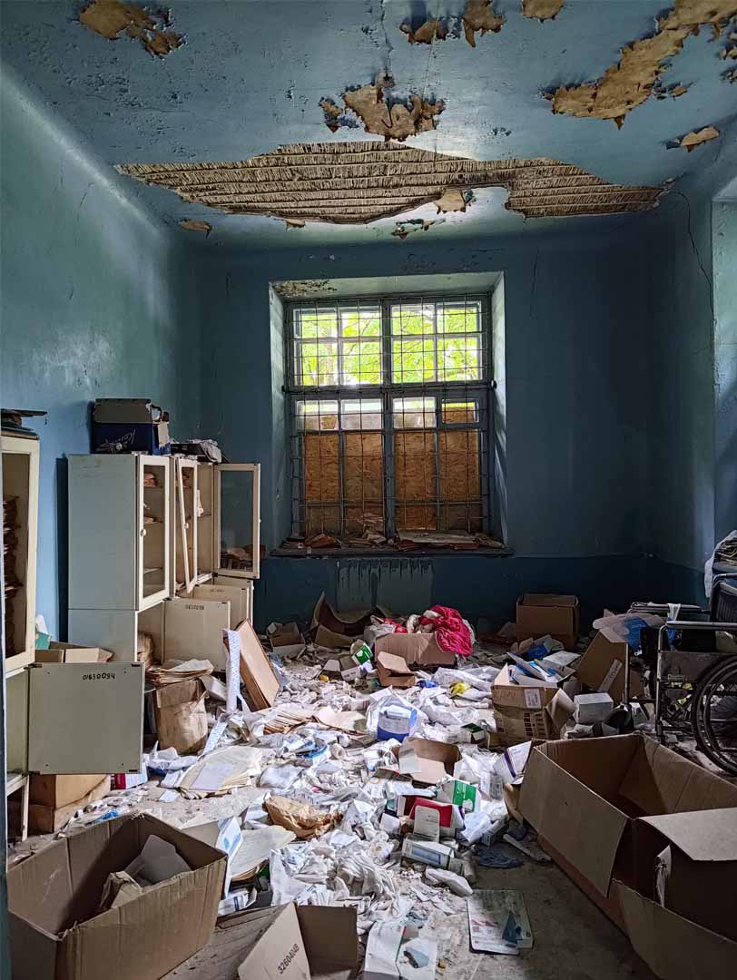 Inside the destroyed medical clinic in Kachkarivka, Ukraine, which is littered with detritus left behind after Russian forces were forced to withdraw in November 2022. The walls are sky blue. The paint in the ceiling is peeled off in some parts. At the center of the photo is a wall with a window that is half boarded up. On the left wall are white cabinets, some have been left half open. The floor is covered in random medical items and medical trash.