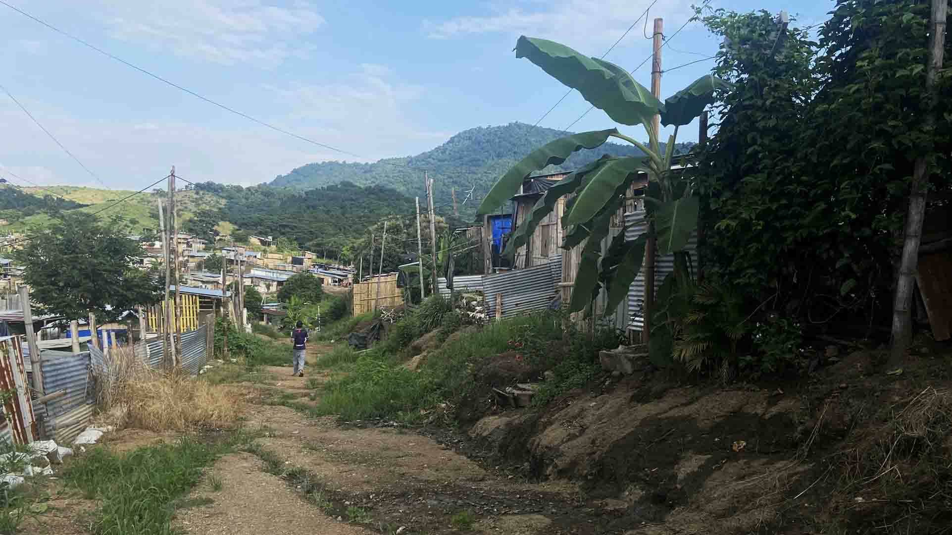 This is a landscape shot of a dirt road in the Nueva Esperanza community. A person is seen walking down the road with their back turned against the camera. Houses are seen in the distance.