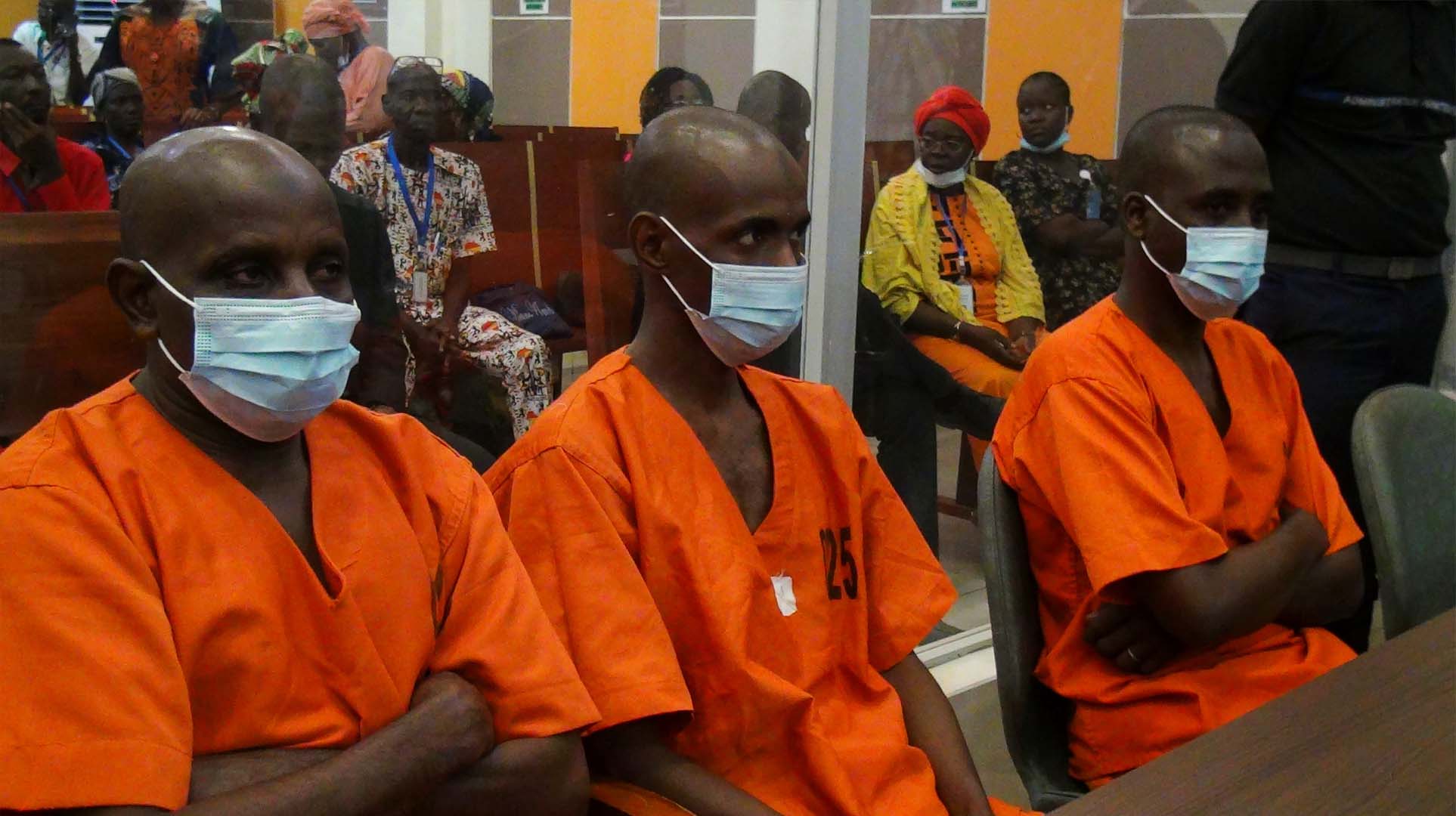 Three men are pictured sitting down, wearing surgical masks and orange prison uniforms.