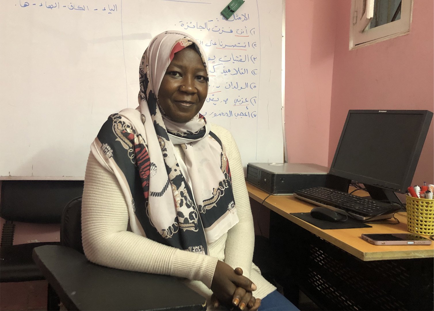 Pictured is Amal Rahal, a 39-year-old architect who left Sudan for Egypt in 2018 and then applied for asylum. She is sat in front of a whiteboard on a black chair. To her left is a desk with a computer.  Her right elbow is on the table and she is smiling at the camera. In 2019, Rahal launched an initiative called Hopes For the Future, with the purpose of setting up a community school to teach Sudanese children in Cairo their own country’s curriculum.