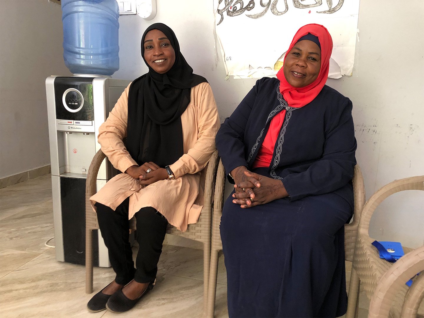 This is a photo of Amany Bubakr. She is pictured sitting on a chair next to a water cooler (to her right) and another woman (to her left) they are both smiling at the camera.