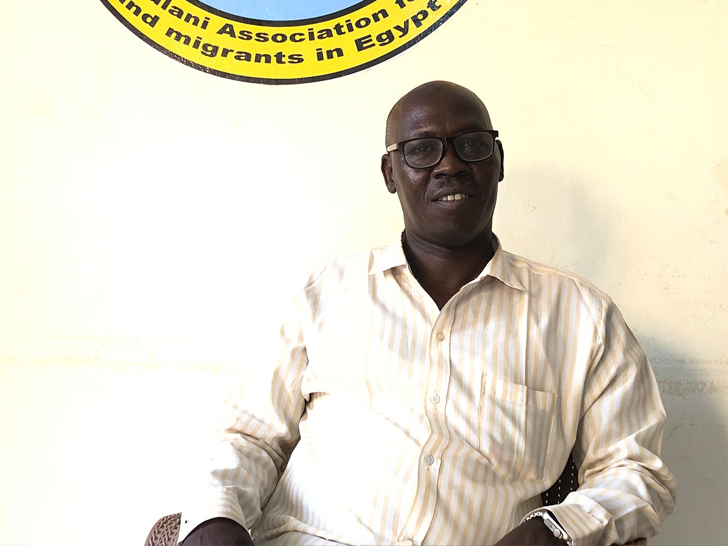 This is a photo of Babiker Tokor, a project leader at the Sudanese Fulani Association for Refugees and Migrants, which was founded in 2016. He is pictured smiling, sitting down in front of a white wall, wearing a button-down shirt and black-rimmed glasses.