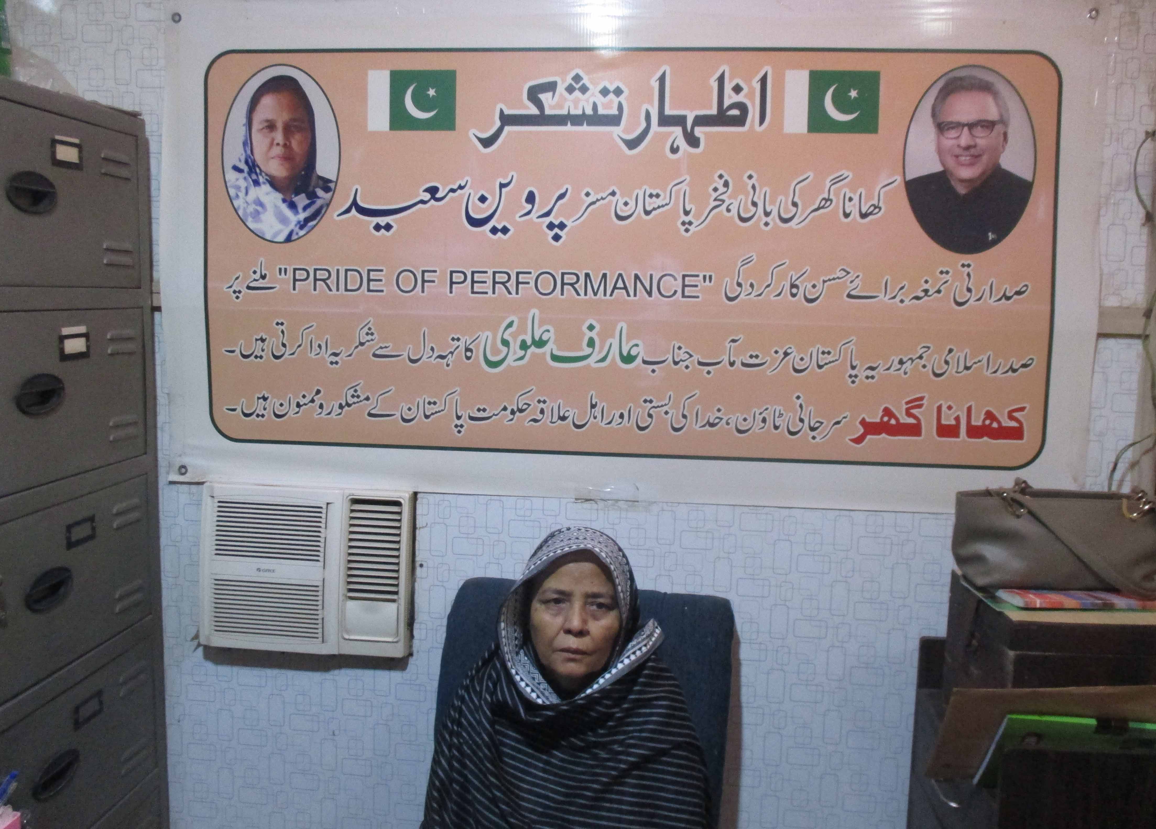 This is a photo of Parveen Saeed, she setup Khana Ghar in 2002. She is pictured under a poster looking at the camera.