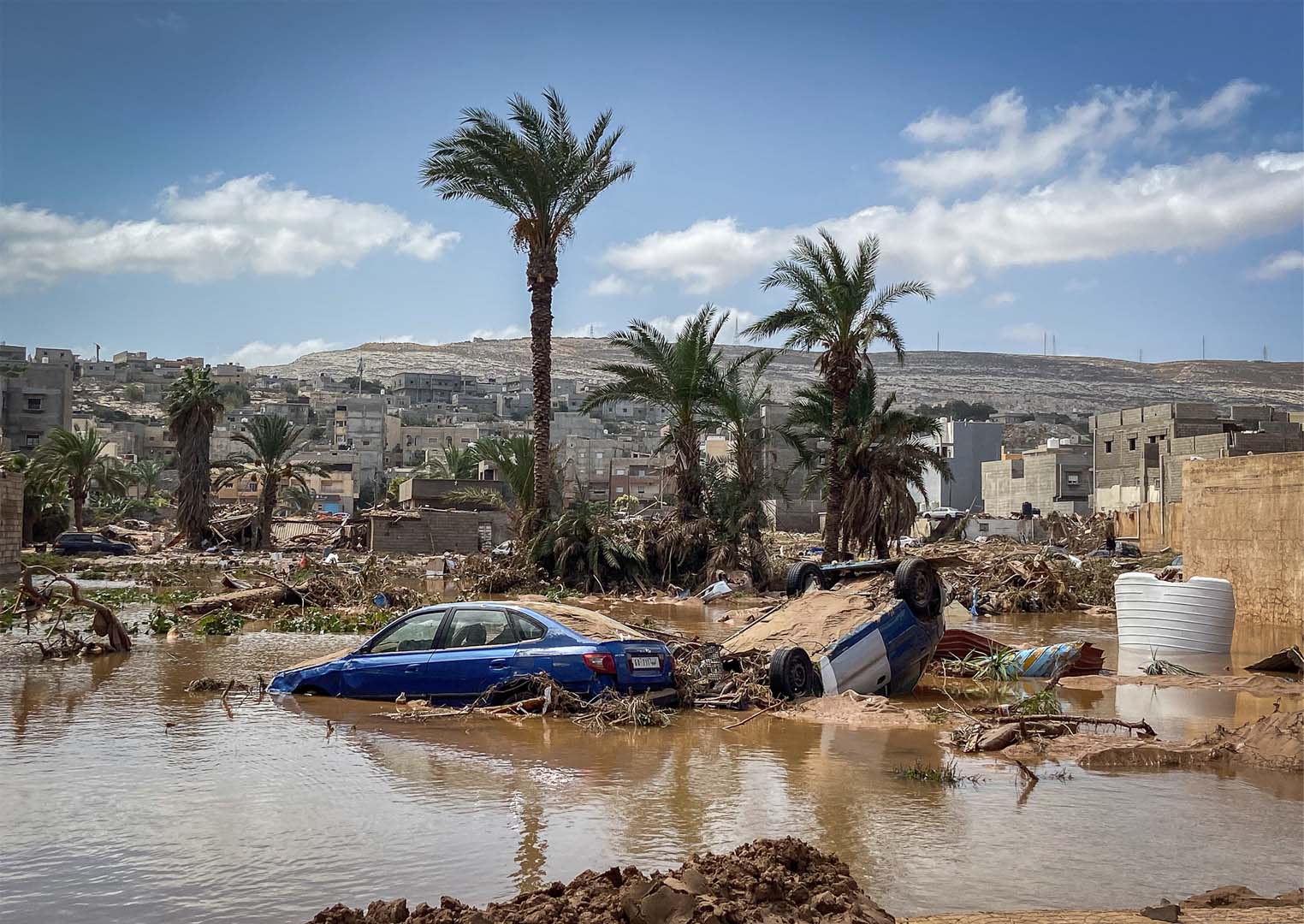 This photo shows the aftermath of the 2023 Libya flood. Pictured in the daytime, there are two cars at the centre of the frame. One is flipped over, mud can be seen on its undercarriage. Another car is next to it, on the left. It’s blue and its wheels are submerged in water. The majority of the lower half of the photo shows water. You can see damaged buildings around and a few palm trees behind the cars.