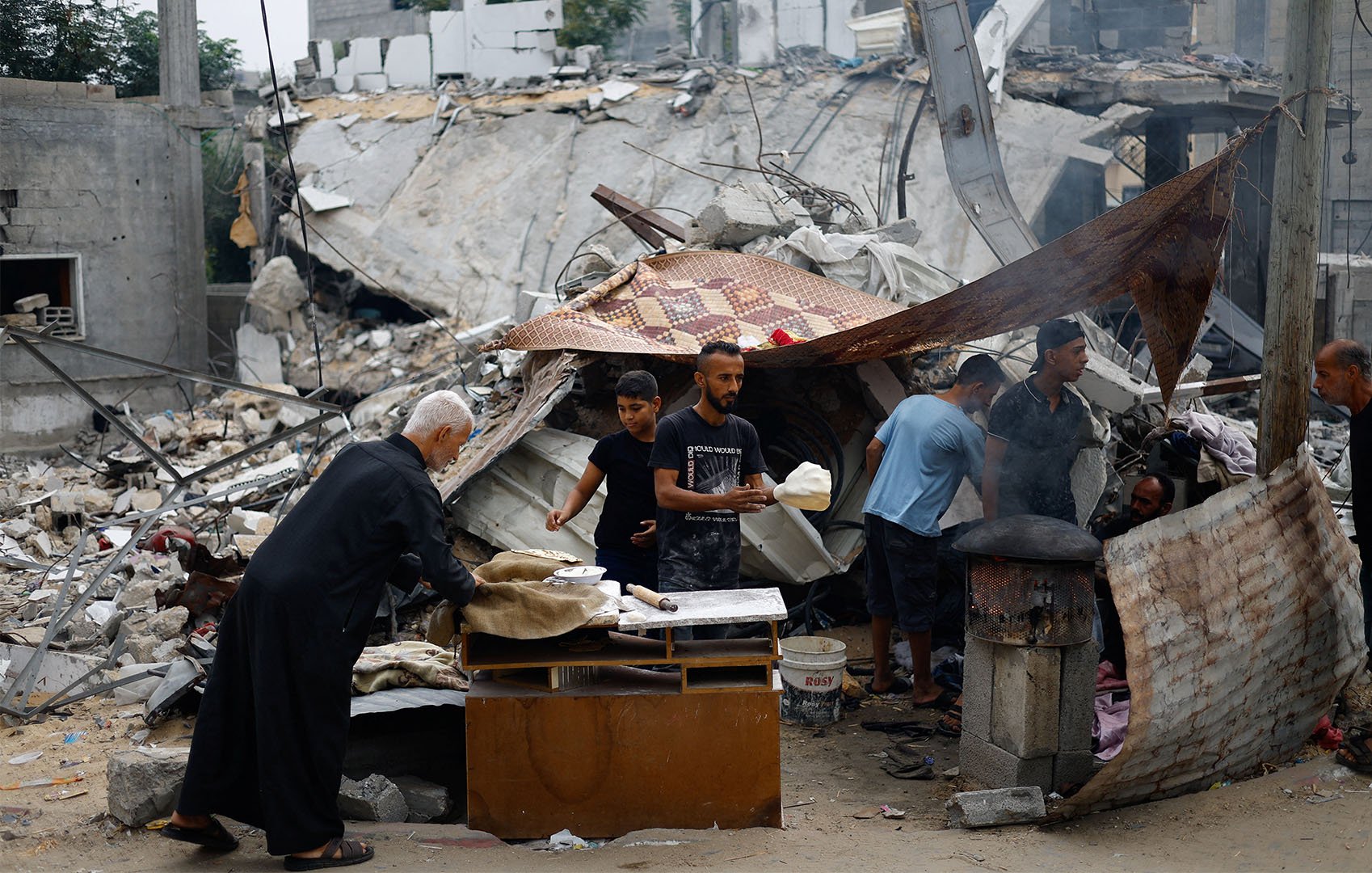 Palestinians prepare food next to destroyed buildings. They are seen cooking in front of rubble on a makeshift fire.