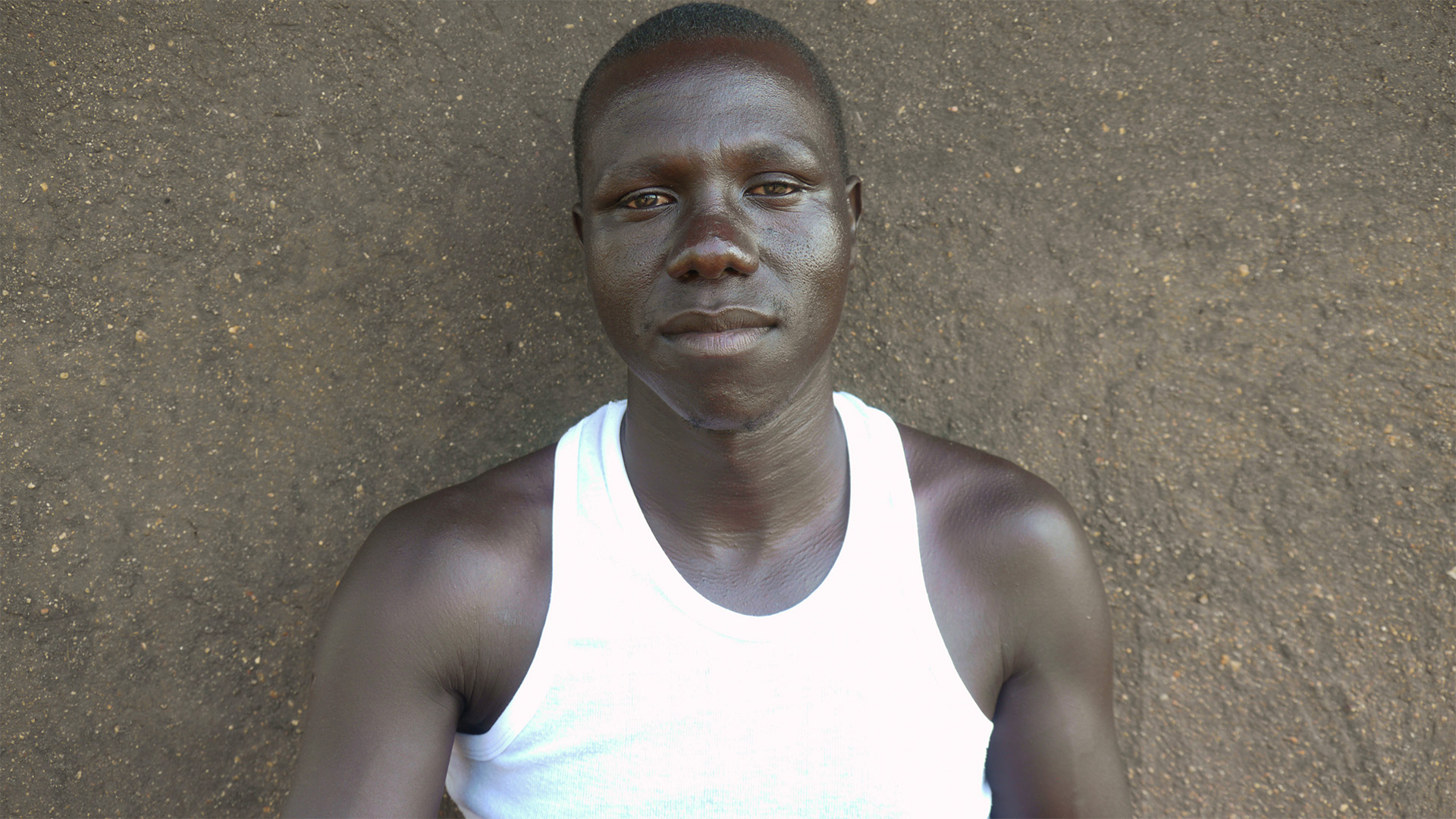 Agwe Lino is pictured wearing a white sleeveless shirt and standing in front of a grey wall.