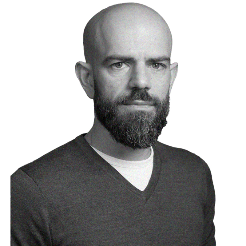 A portrait image of Tammam Aloudat in black and white. He looks at the camera and smiles without showing teeth. He wears a dark v-neck sweater