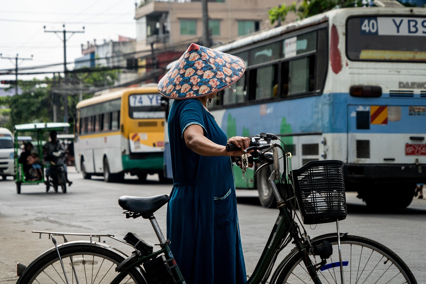 A person wearing a floral hat walks through a street market while holding on to a bicycle. 