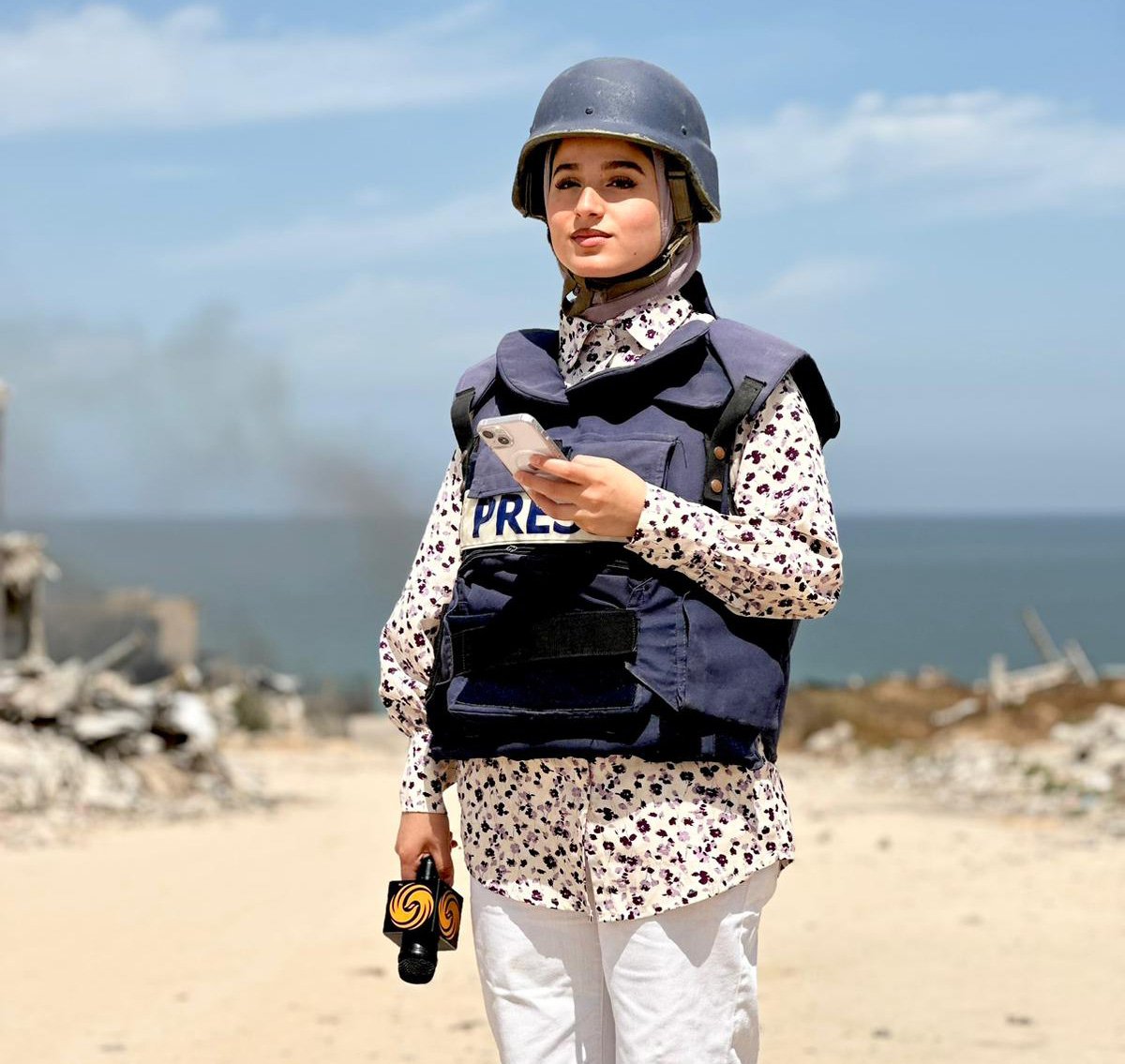 A photo of Palestinian journalist, Nour ElAssy. She wears a press vest and helmet. She holds a phone with her left hand and a microphone with her right hand.
