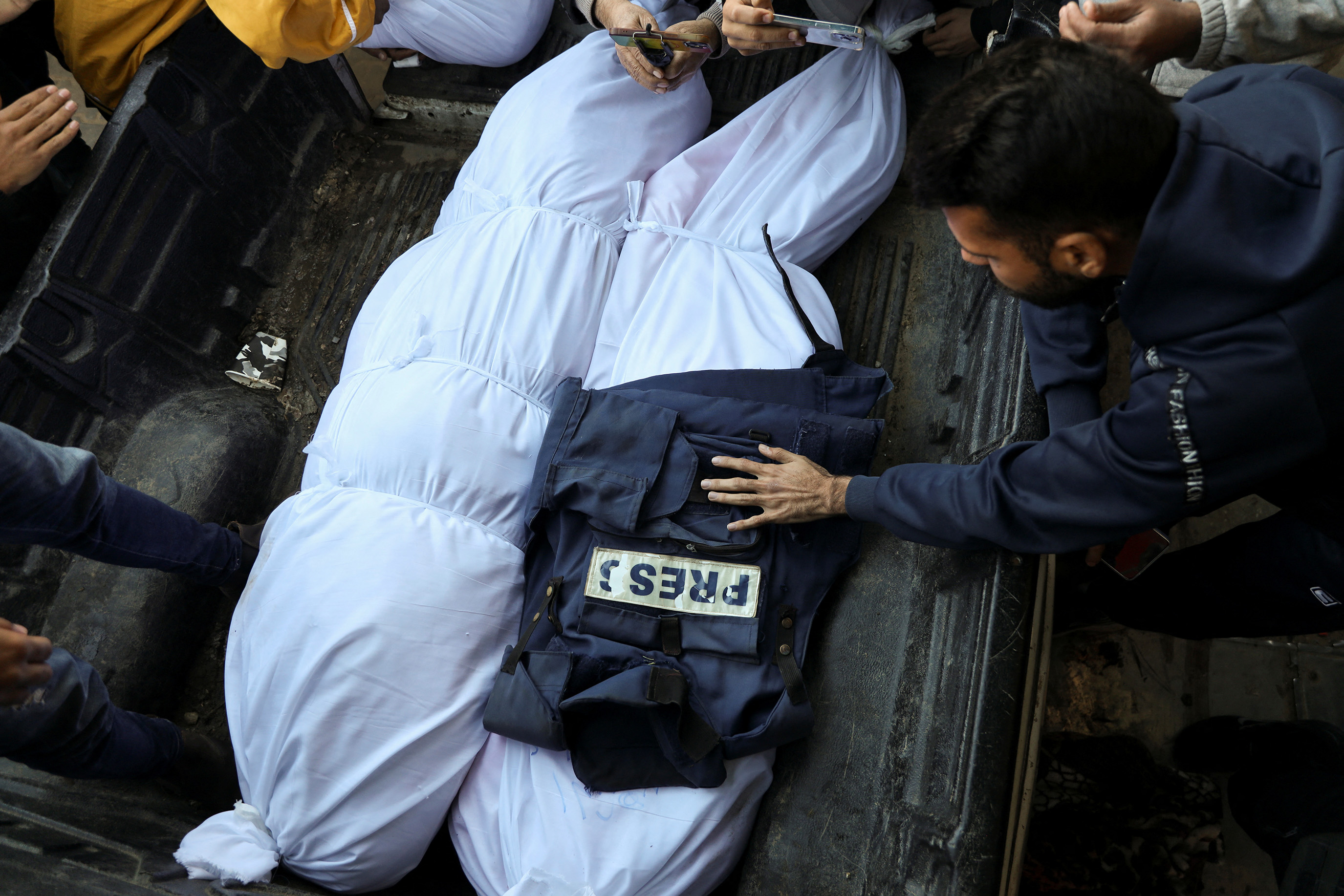 A photo of mourners as they gather around the bodies of two Palestinian journalists who were killed in Israeli strikes, amid the Israel-Hamas conflict. Their bodies are wrapped in white sheets and one has a press vest on it.