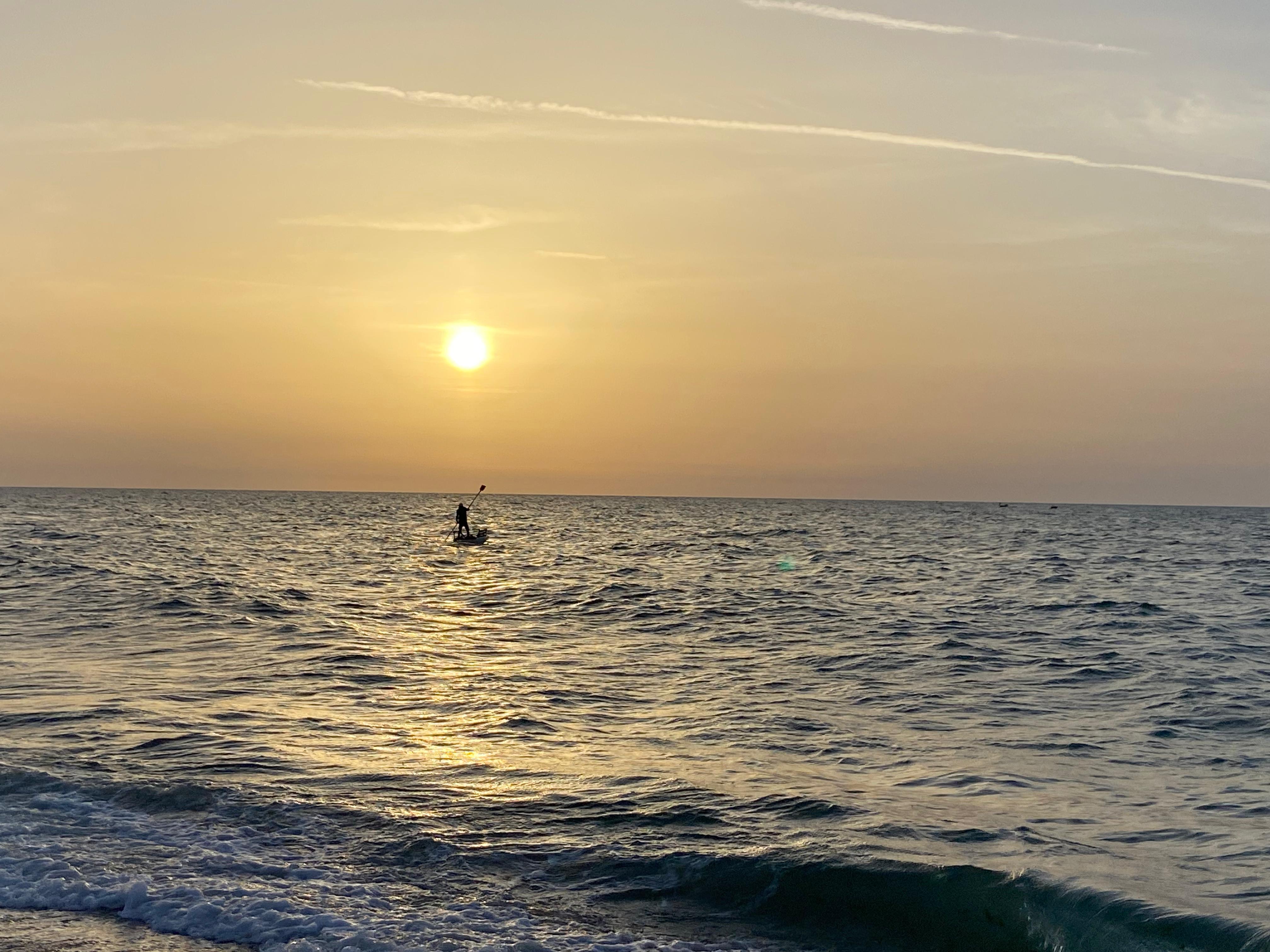 A photo of the sea on Rita’s last visit, the day before she left Gaza. A person can be seen rowing in the water as the sun sets behind.
