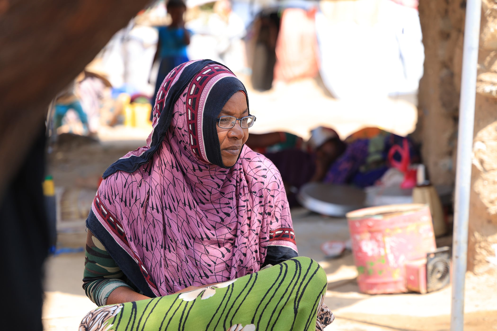 A portrait of Hafedhaa Murshed. She wears a pink patterned headscarf and glasses as she sits and looks to the left.
