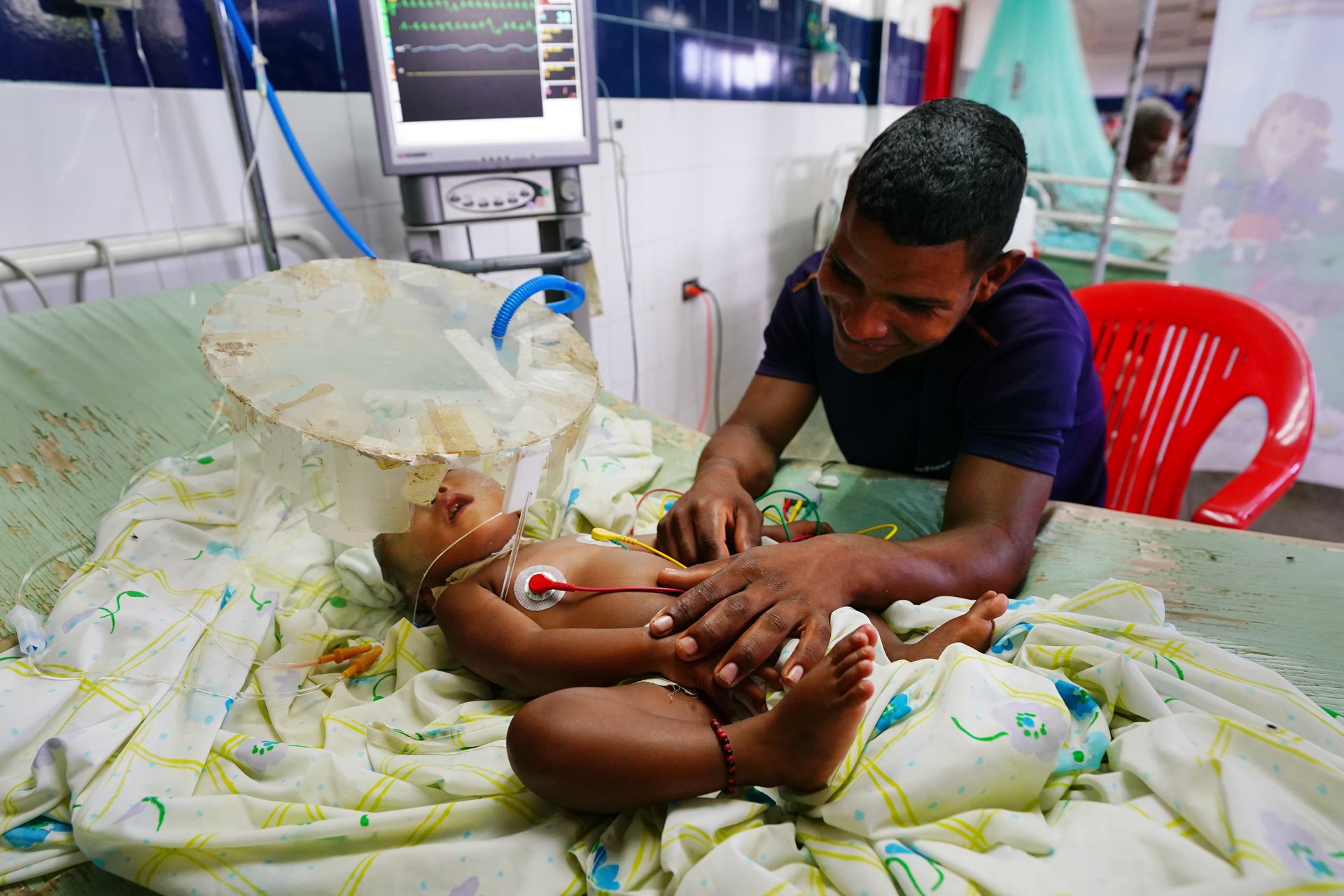 A man over an infant in a hospital bed connected to tubes