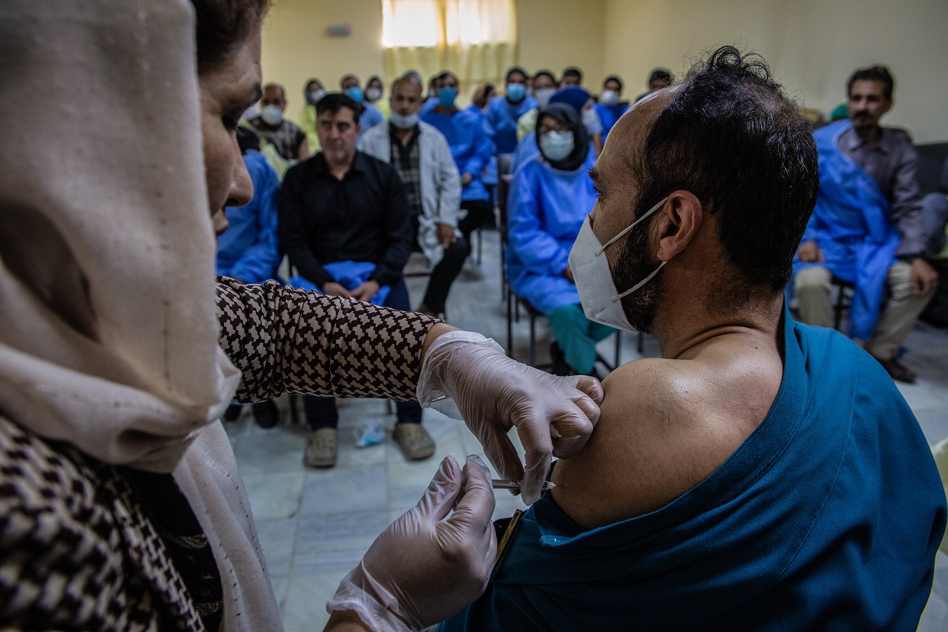 A nurse gives a vaccine shot in a man's arm in front of many patients.