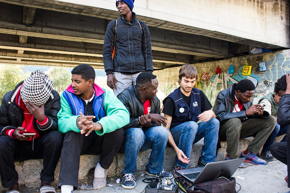 Asylum seekers and a volunteer sit outside around a laptop and equipment