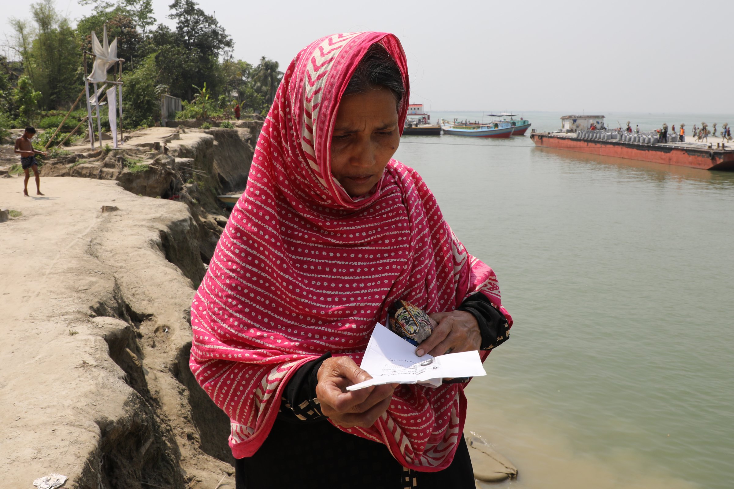 A woman stands on the bank of a river in Bangladesh reading a small piece of paper