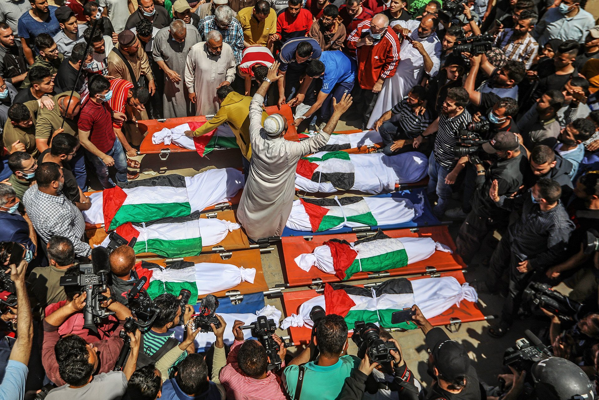 A crowd of people gather around several bodies wrapped in Palestinian flags.