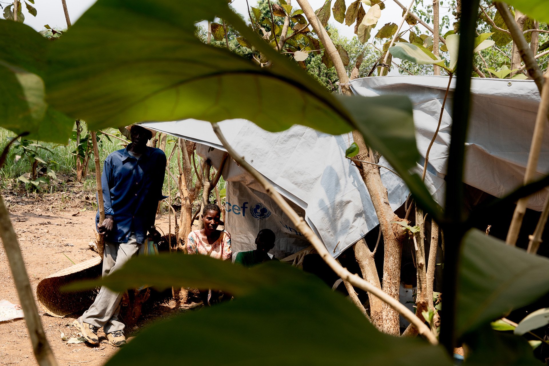 A man stands beneath a makeshift shelter, his face half shaded by the shadow cast by the tarp. 