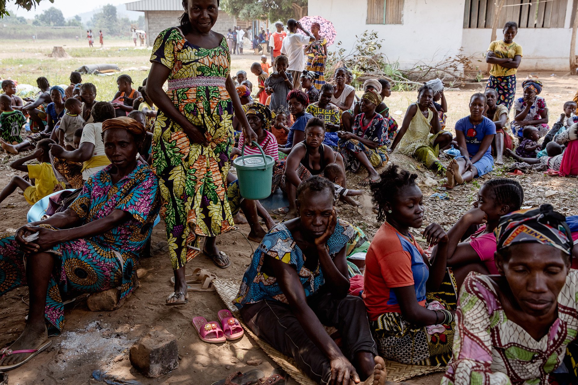 A group of villages of all ages fill the frame, sitting on the ground in a displacement camp. 