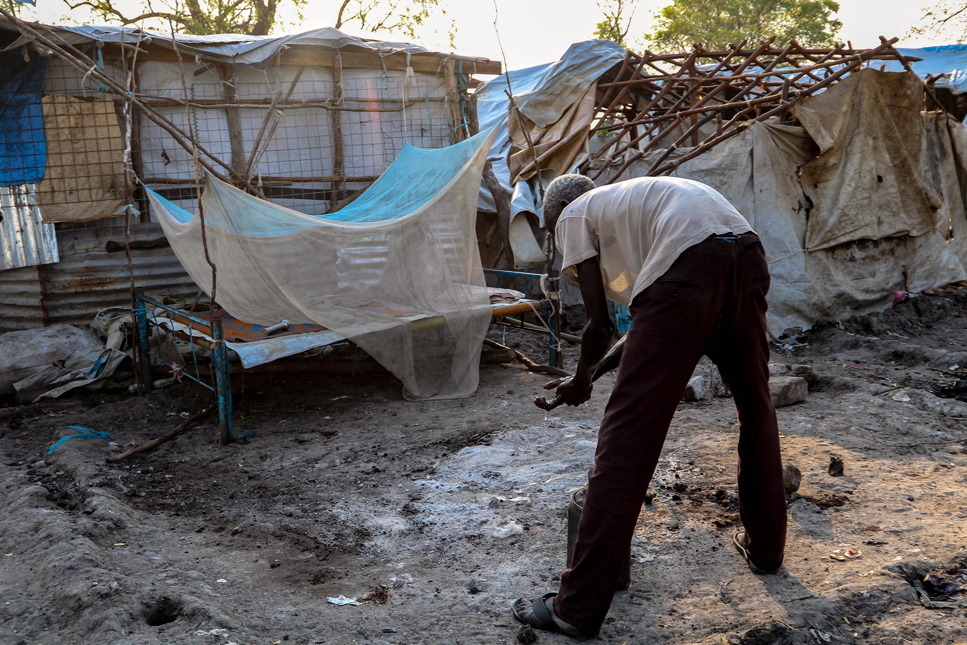 A displaced man with no shelter bends over a shallow bucket in a clearing between makeshift shelters.