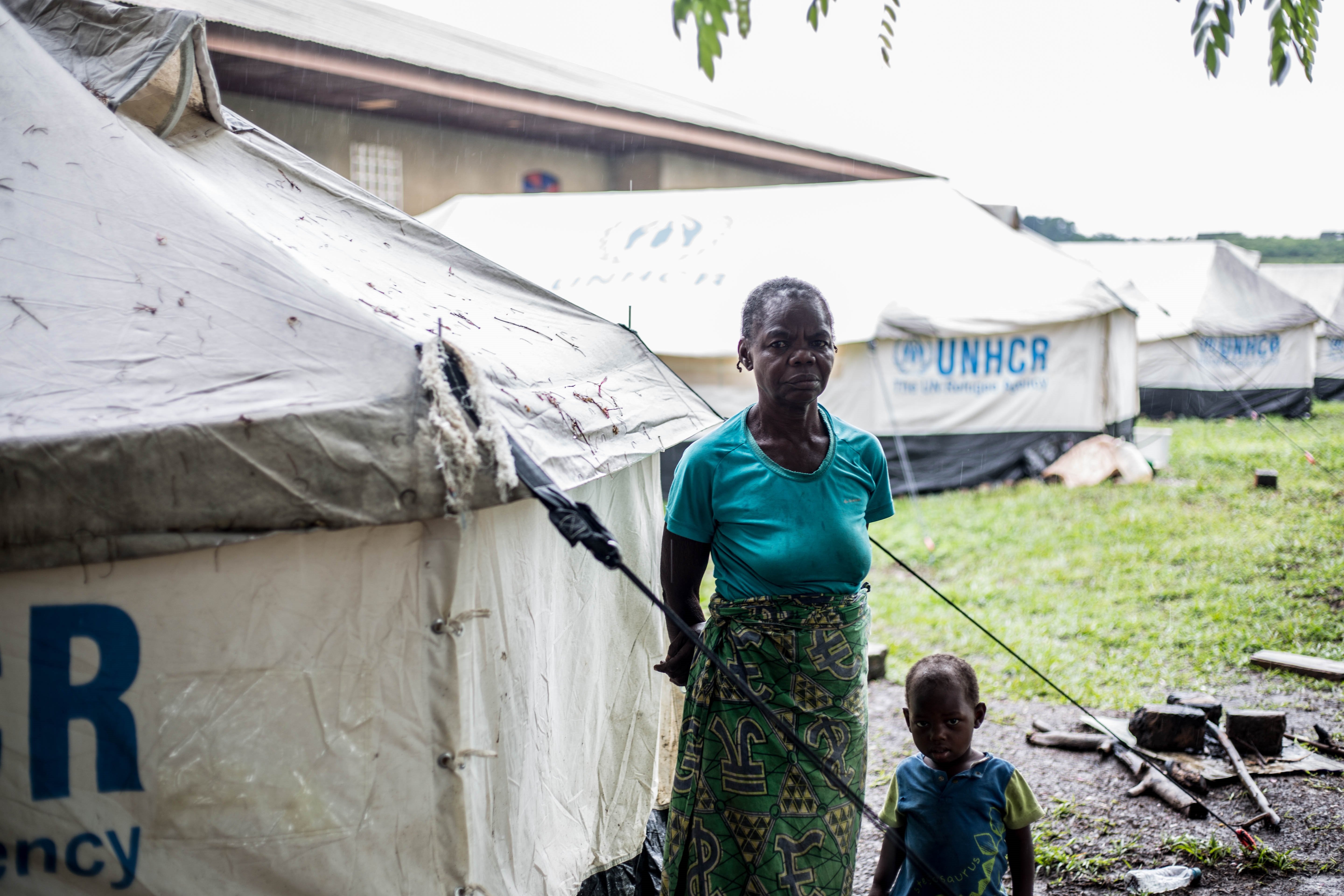 A woman and a child stand in front of a UNHCR tent in the rain facing the camera