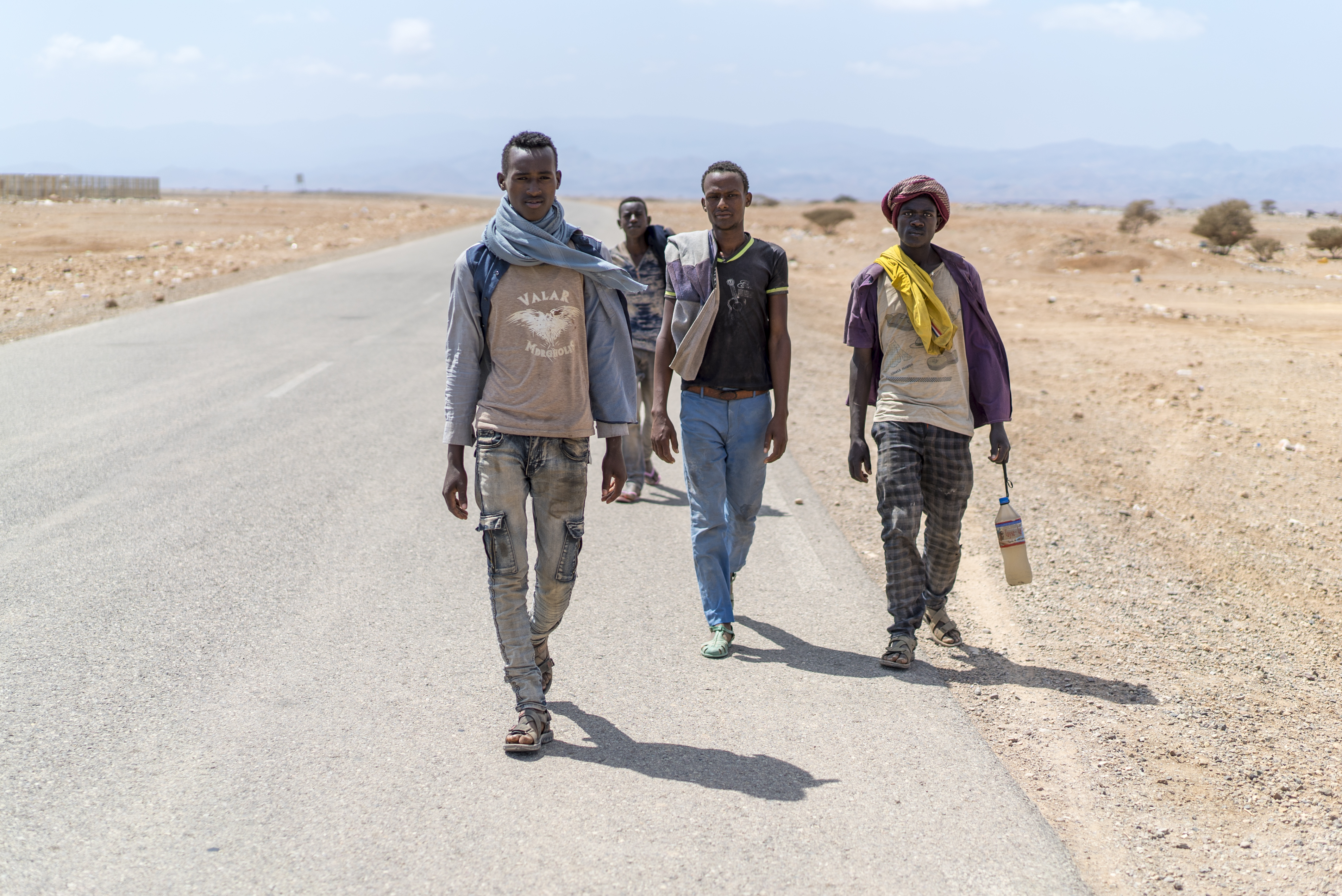 Four young men walk toward the camera on a road in an arid landscape with little else surrounding them