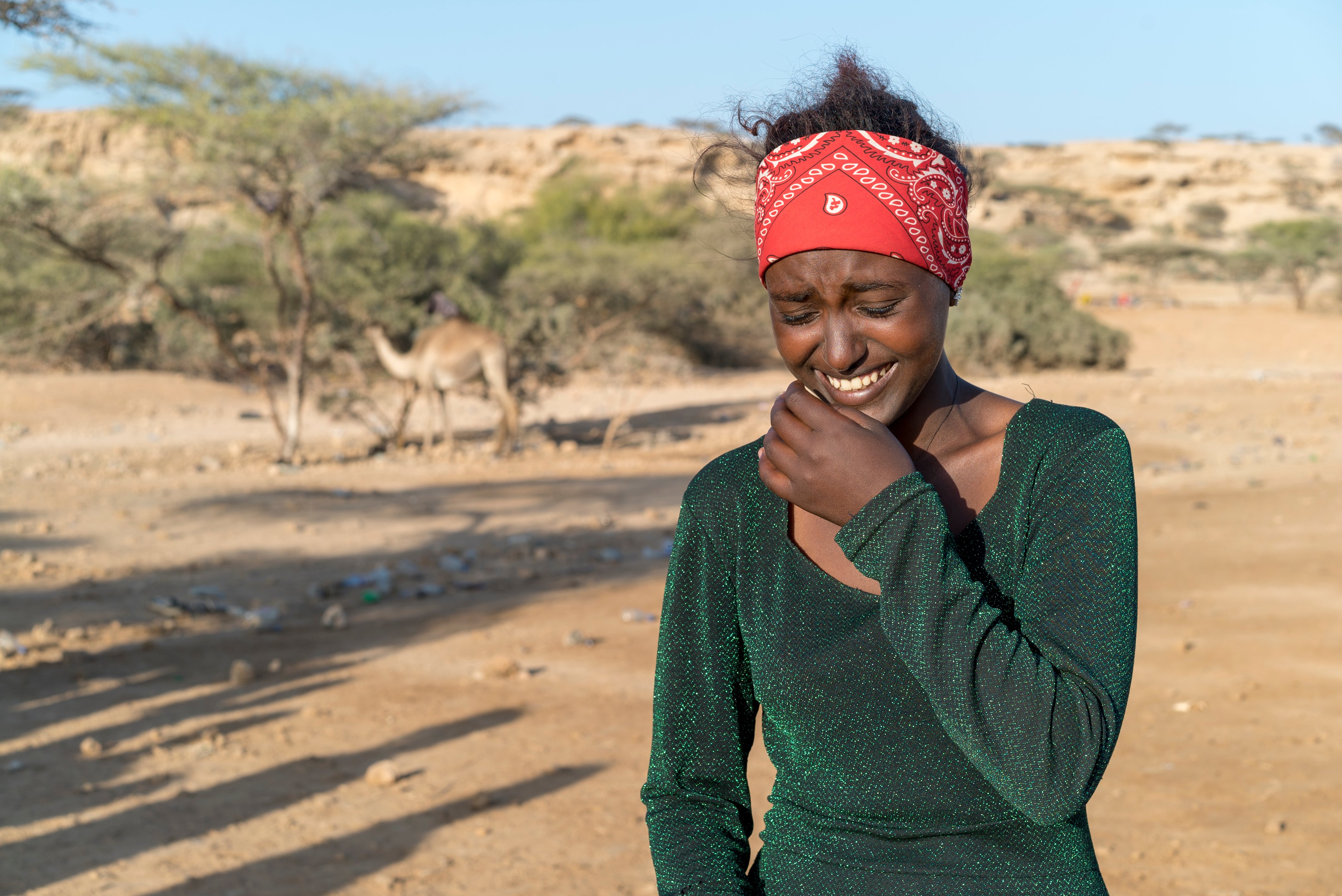 A woman with a green long sleeve shirt and a red bandana in her hair laughs. There is a camel in the background 