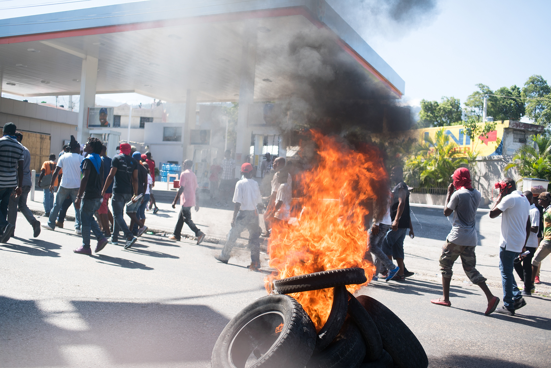 A stack of tires on fire at a petrol station as protesters march behind