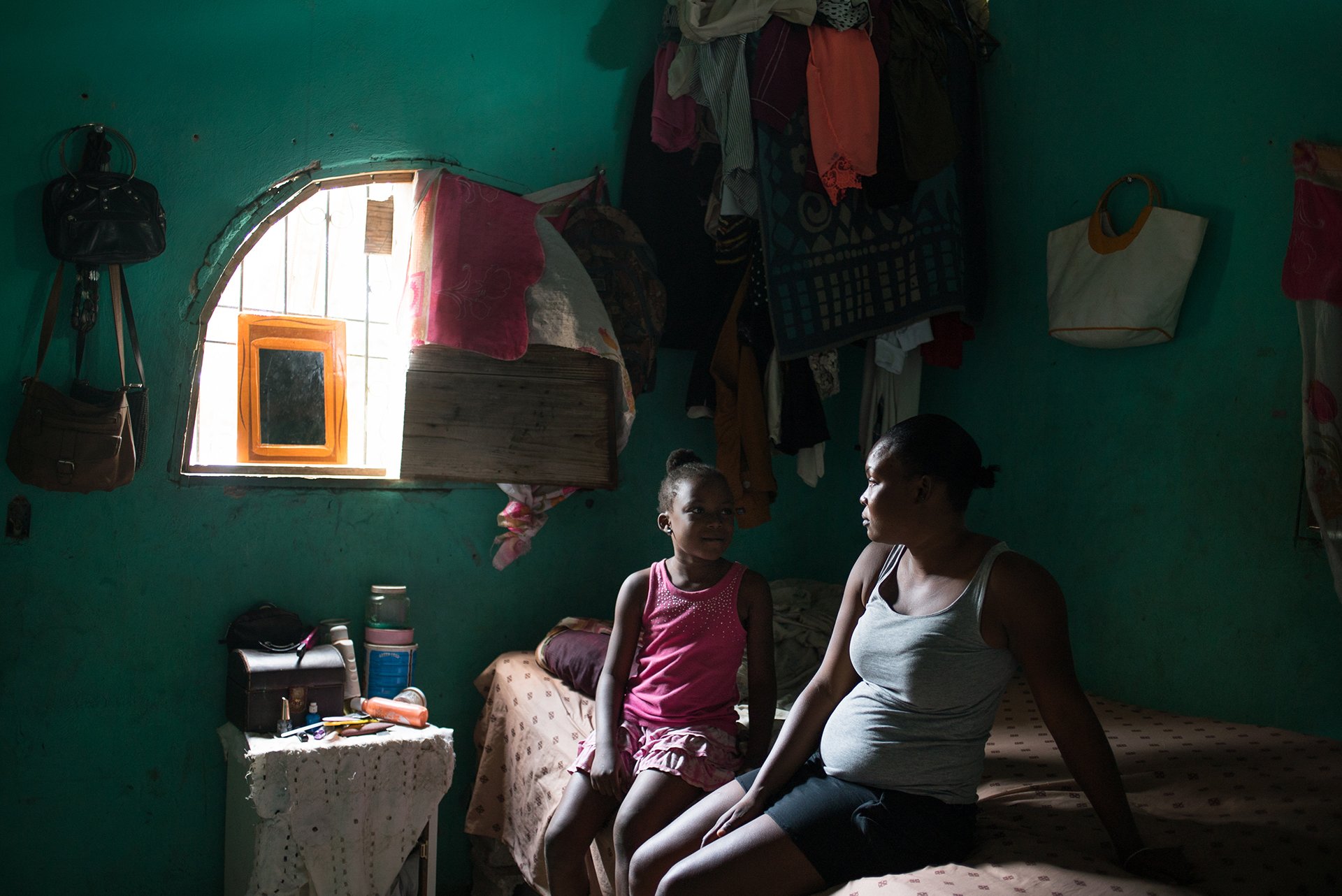 A mother and daughter sit on a bed as light from the window falls on them