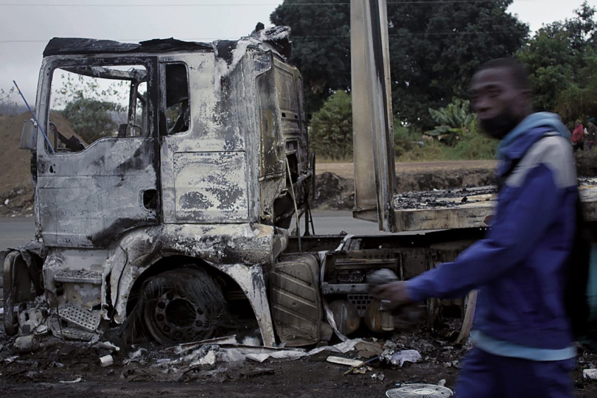 A man walks in front of a burnt out truck. 