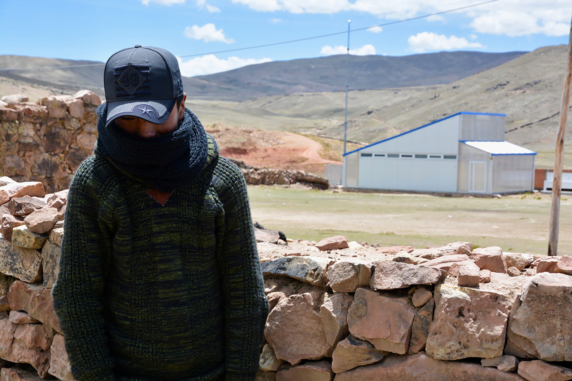 A boy, his face covered by the brim of his hat, stands facing the camera in a rural location.