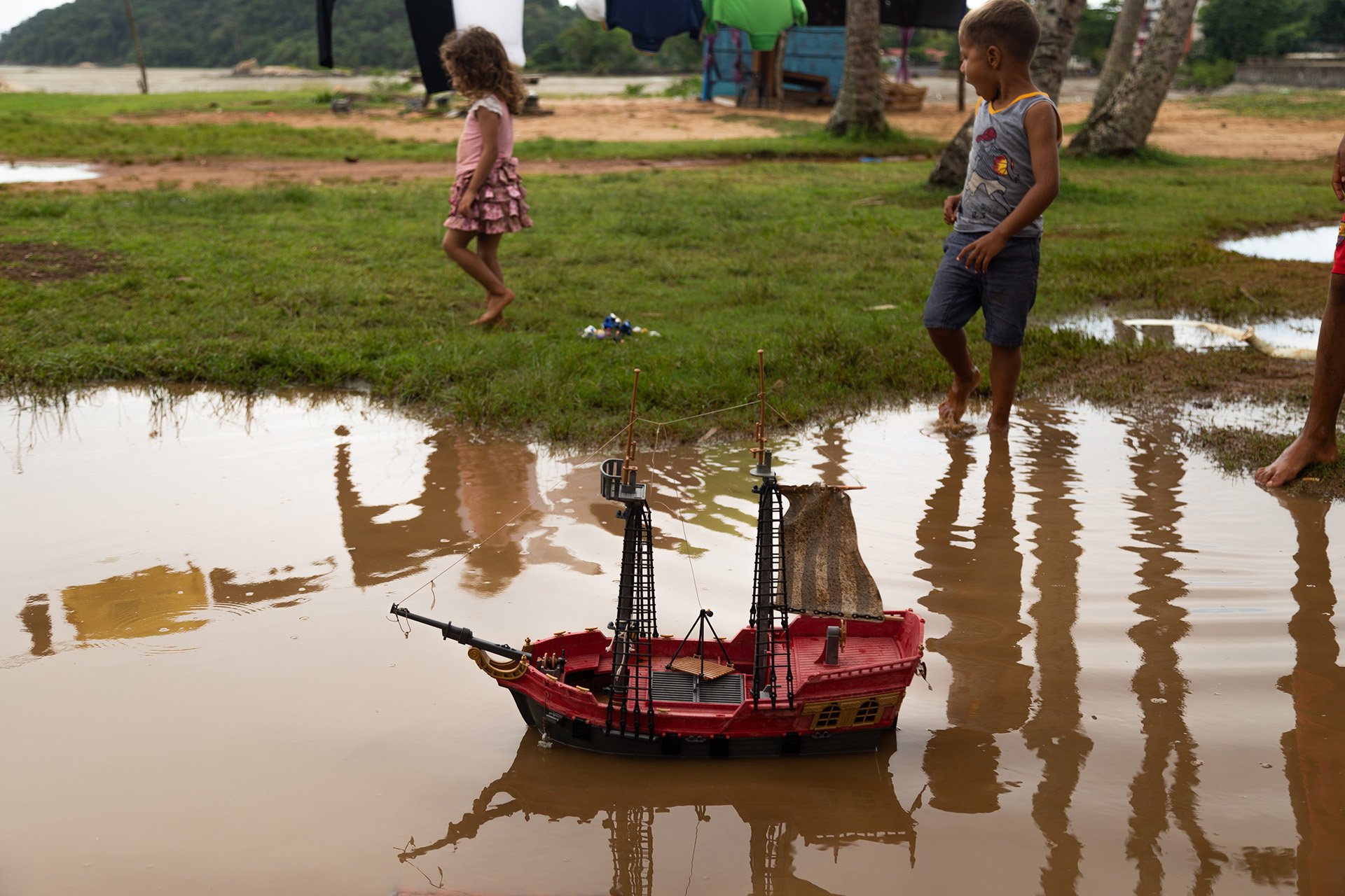 Two young children walk by a toy boat as it floats in a shallow puddle. 