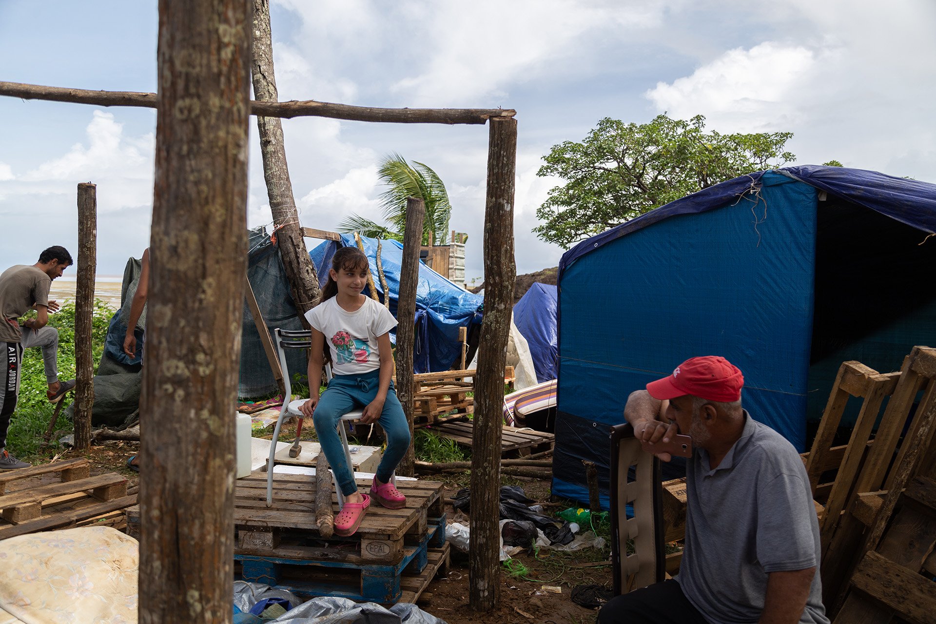 A young girl sits on a pile of wood speaking with an older male relative. 