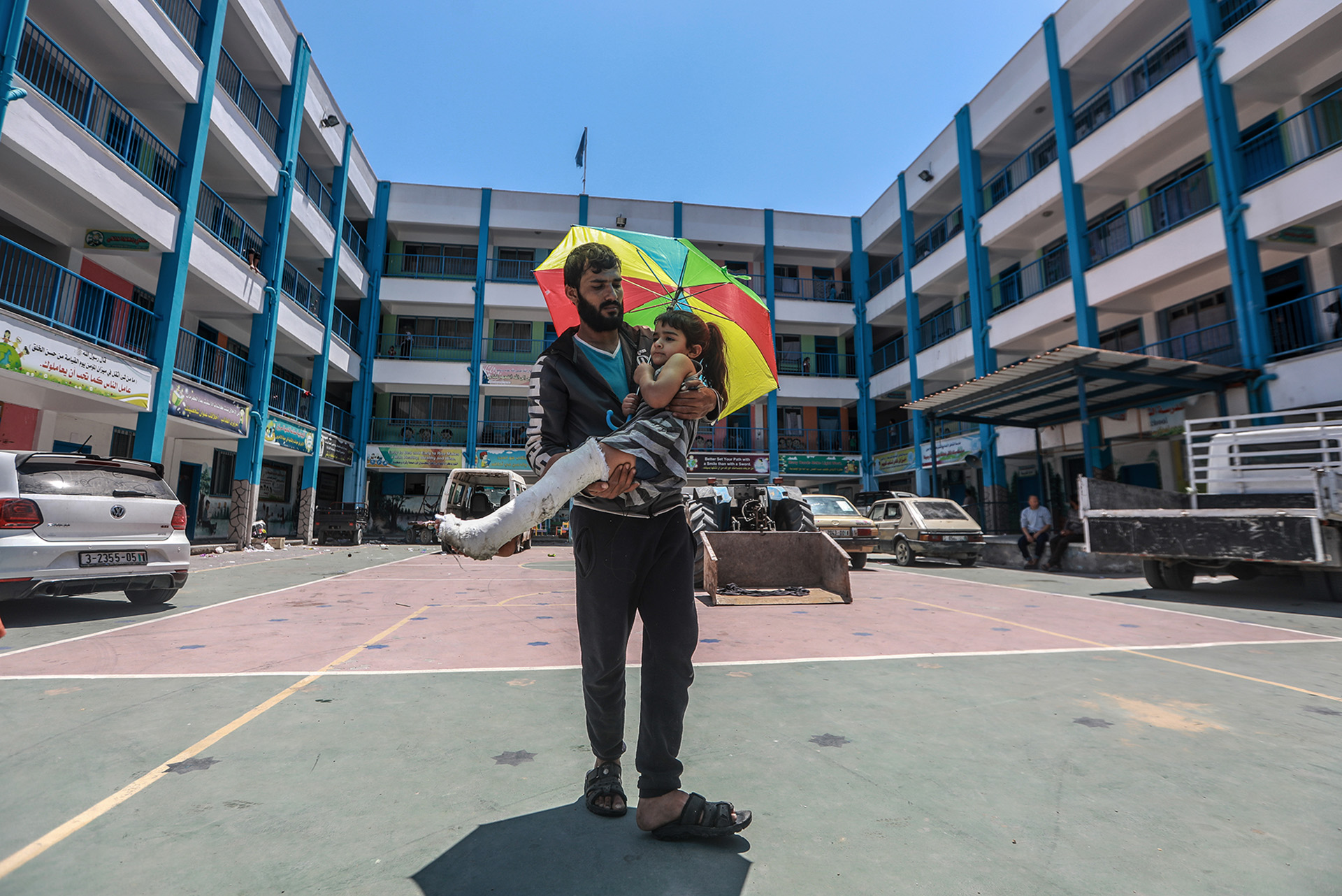 A girl with a bandaged leg holding a multi-colored umbrella is carried by a man outside of a hospital. 