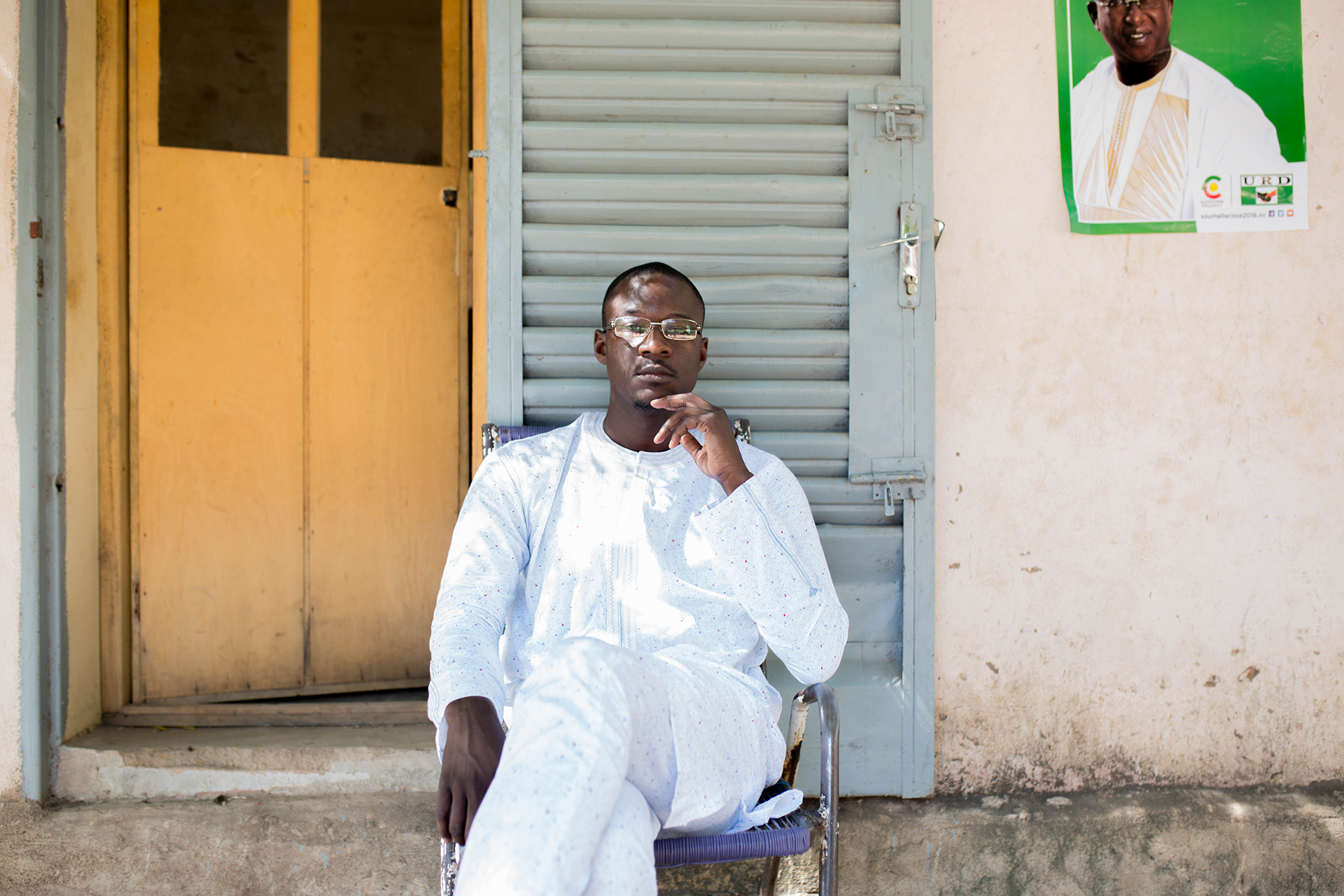 A man in white looks directly into camera sitting in front of a building wall.